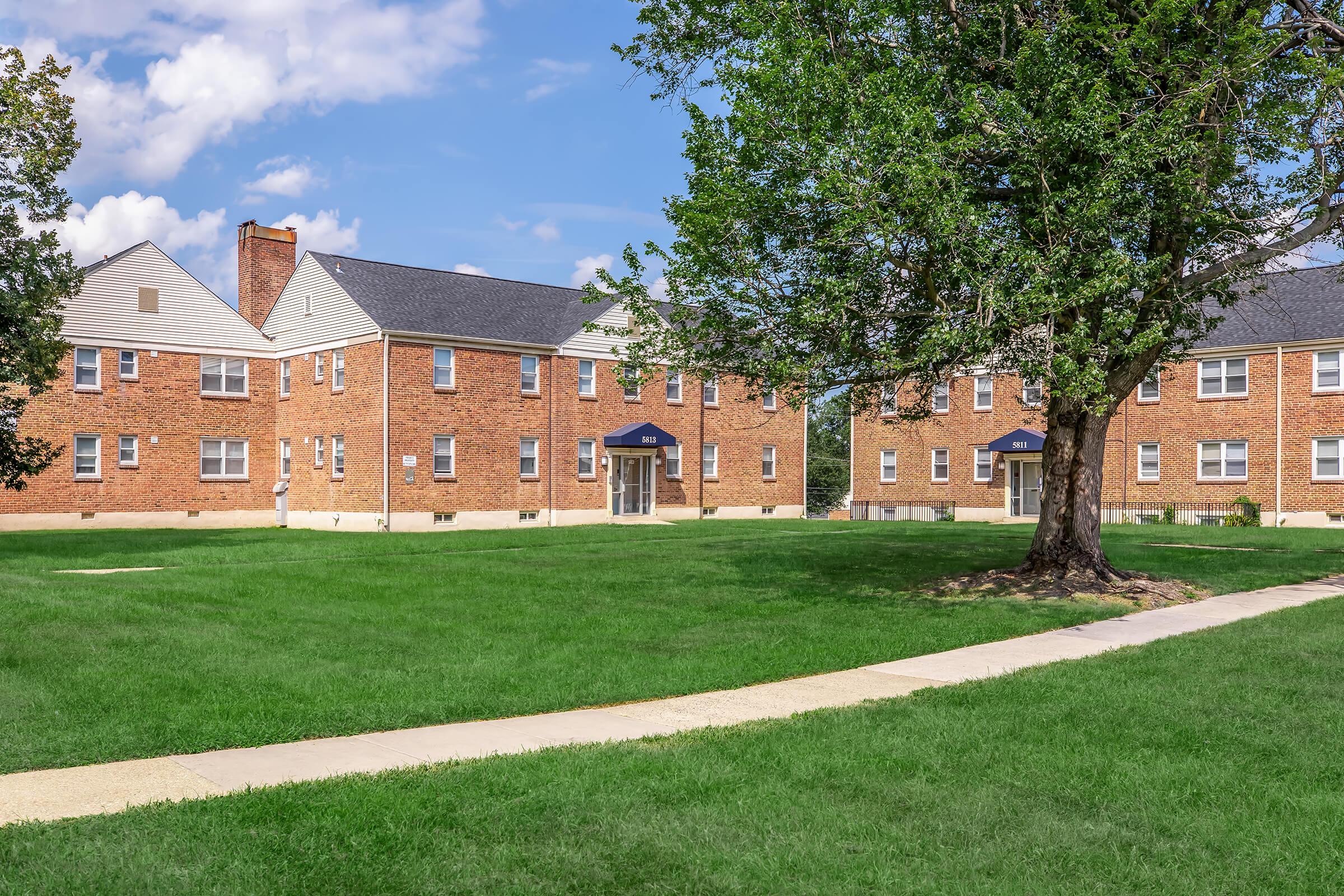 A well-maintained brick apartment complex featuring multiple buildings, surrounded by a green lawn and mature trees. A paved walkway leads through the grass, connecting the buildings. The sky is partly cloudy, creating a bright and inviting atmosphere.
