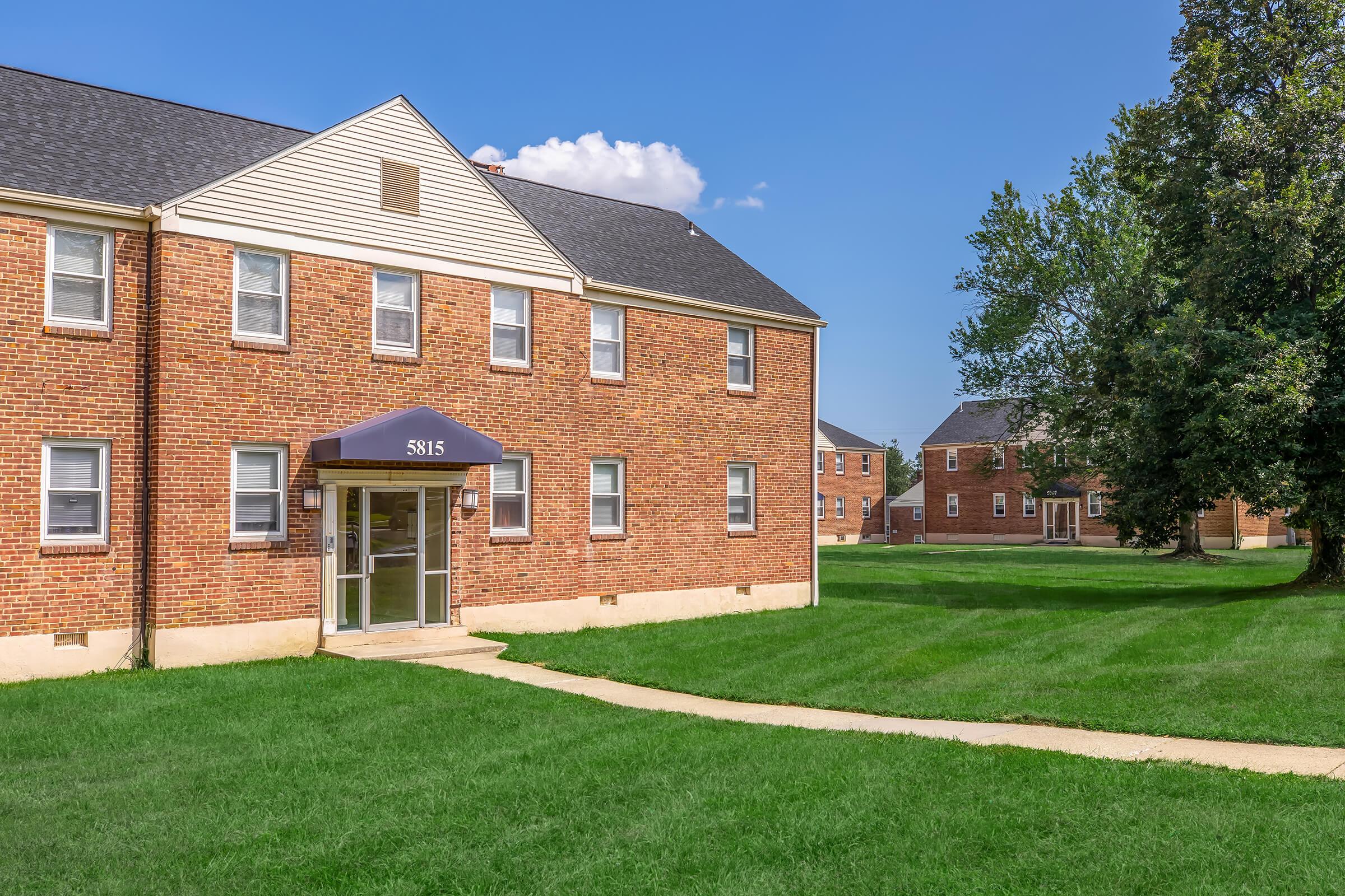Brick apartment building with a dark awning over the entrance. The setting includes a well-maintained lawn with a concrete path leading to the entrance. In the background, additional residential buildings and trees are visible under a clear blue sky.