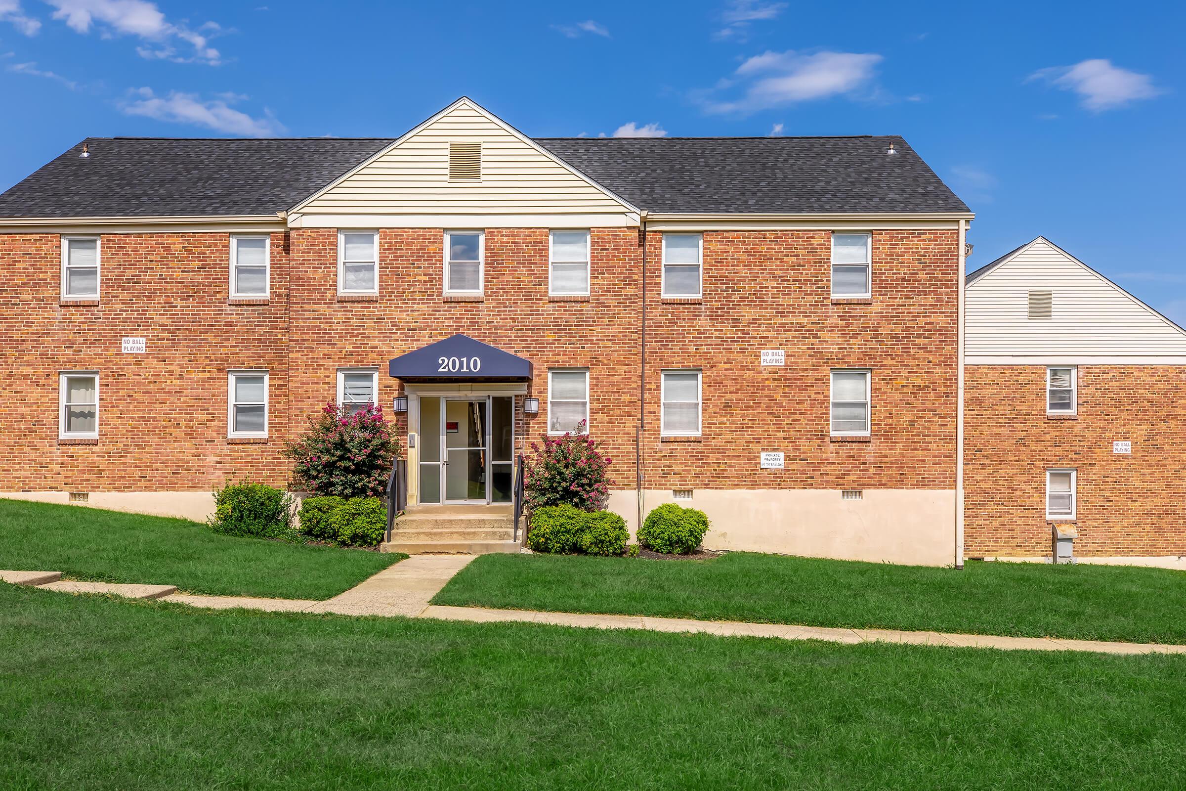 Brick apartment building with white trim and a dark blue awning, featuring multiple windows. The entrance is marked with the number "2010." Lush green grass surrounds the walkway, and there are small bushes and flowers near the building. The sky is clear with a few clouds.