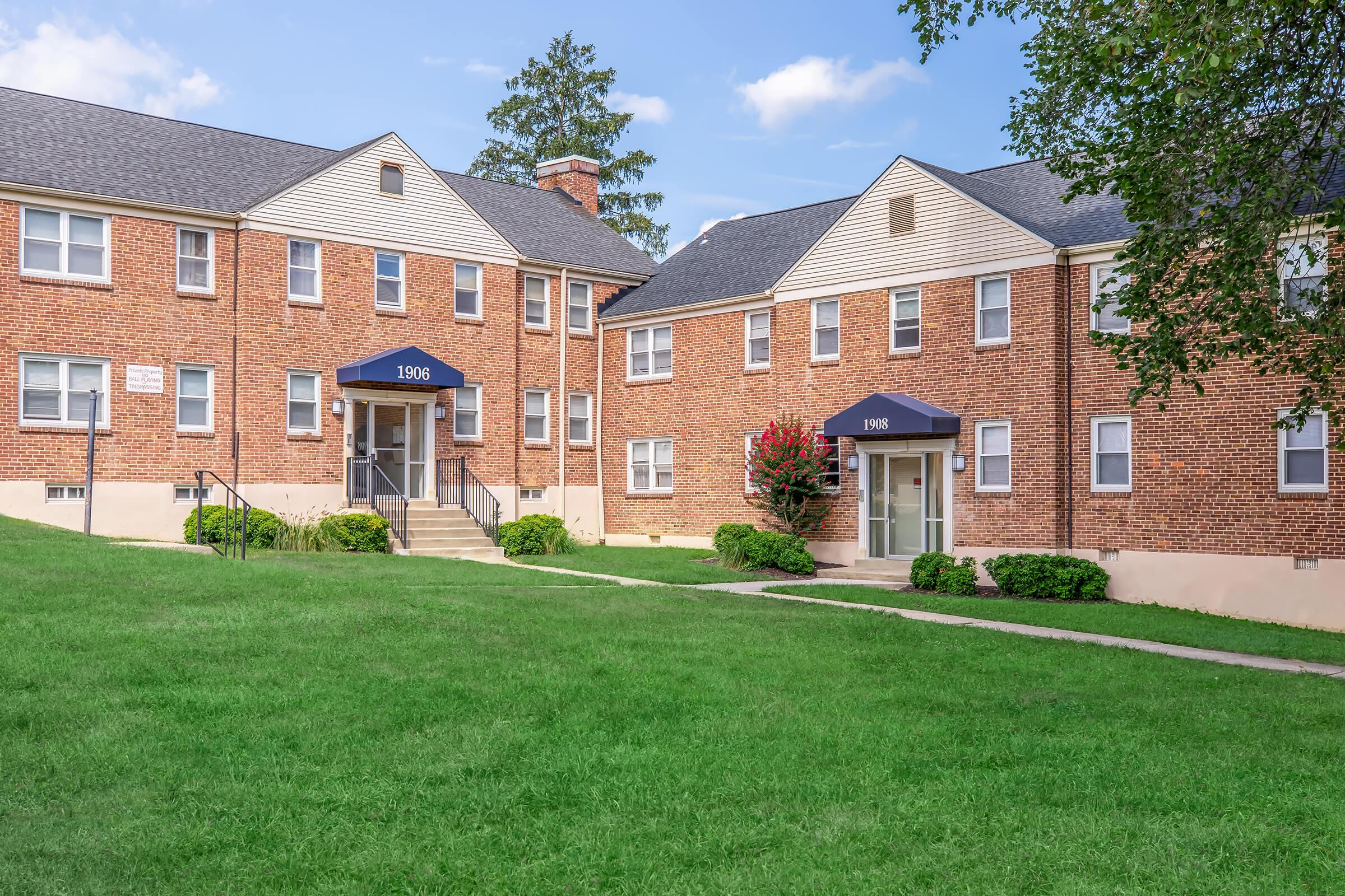 Two brick apartment buildings with flat roofs, labeled 1096, situated on a grassy area. Each building has a set of stairs leading up to the entrance, with green shrubs and trees nearby. The sky is clear with a few clouds, creating a serene residential atmosphere.
