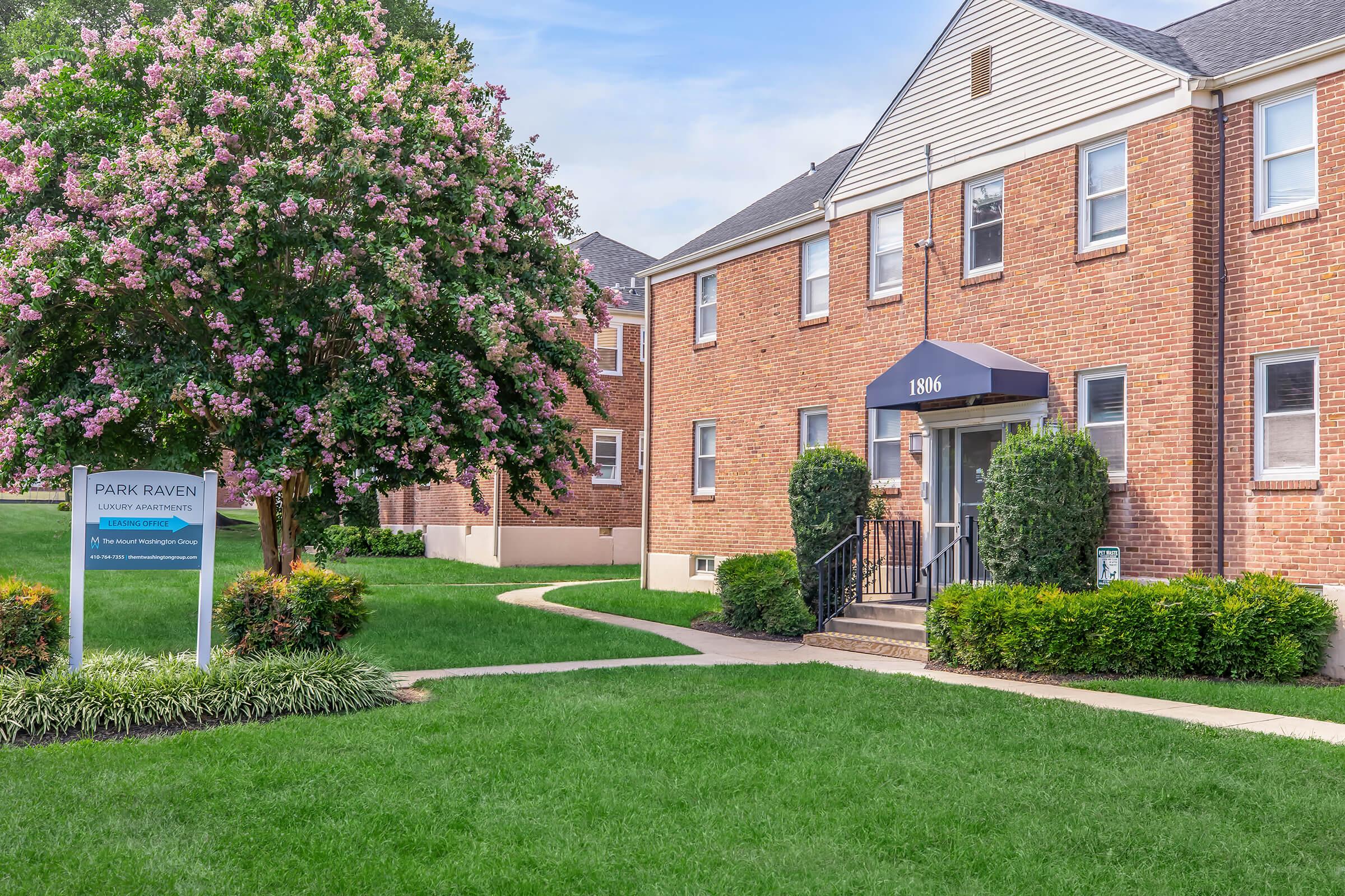 A brick apartment building with a dark blue awning at the entrance, surrounded by green lawns and shrubs. A large flowering tree provides shade nearby. A sign for "Park Raven" is prominently displayed at the front, indicating the location of the rental community. Clear blue skies above.