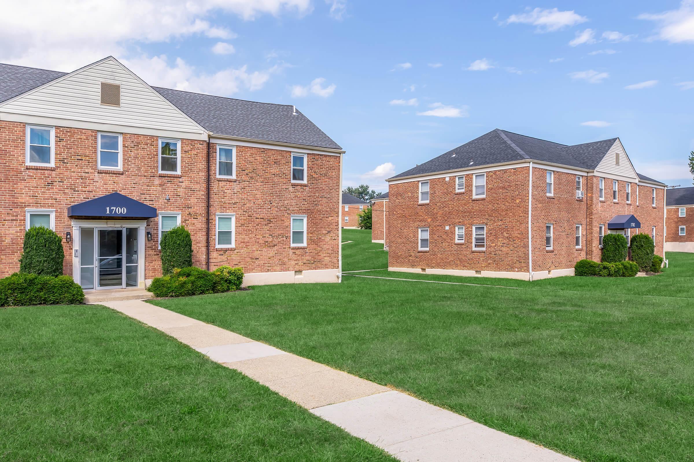 Brick apartment buildings with blue awnings and green lawns, featuring a clear sky in the background. The walkway leads to the entrance of one building, while another building is visible in the distance, both surrounded by well-maintained grass.