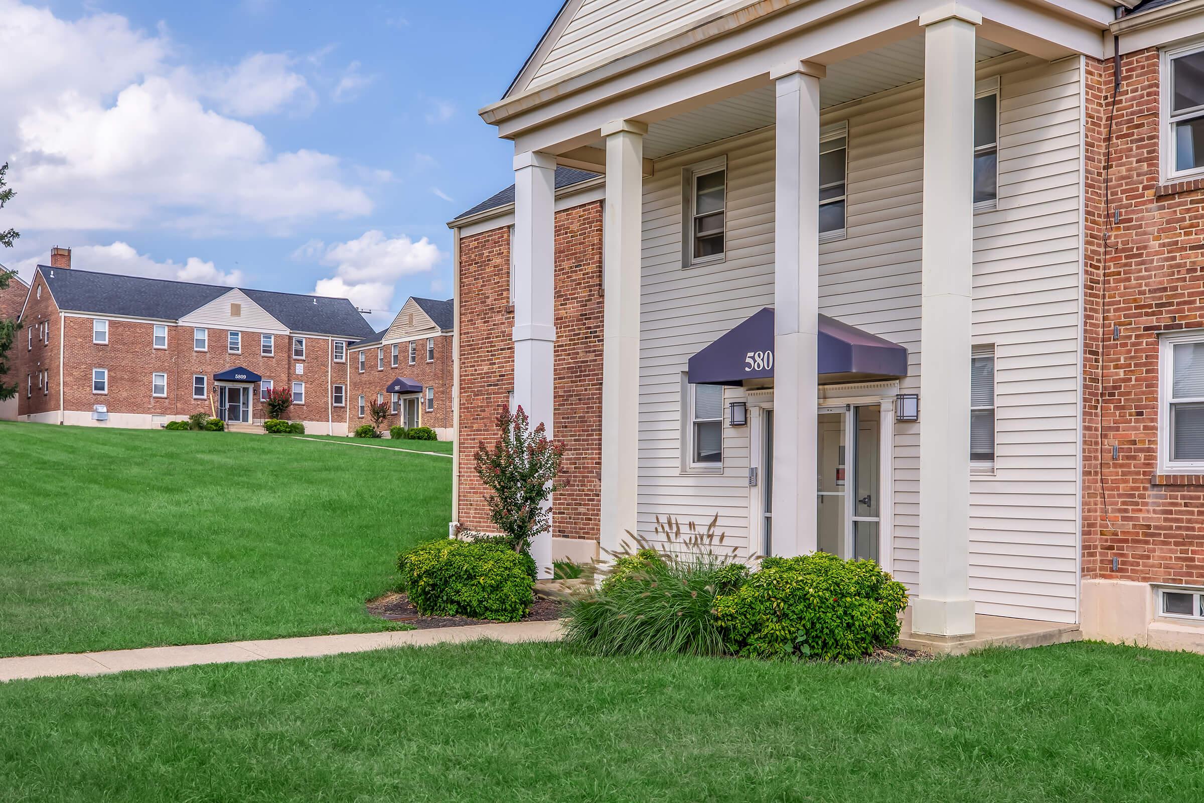 A brick apartment building with white columns and a gray awning at the entrance, surrounded by a well-manicured lawn. In the background, additional brick buildings are visible, and the sky is partly cloudy. The scene conveys a residential neighborhood atmosphere.
