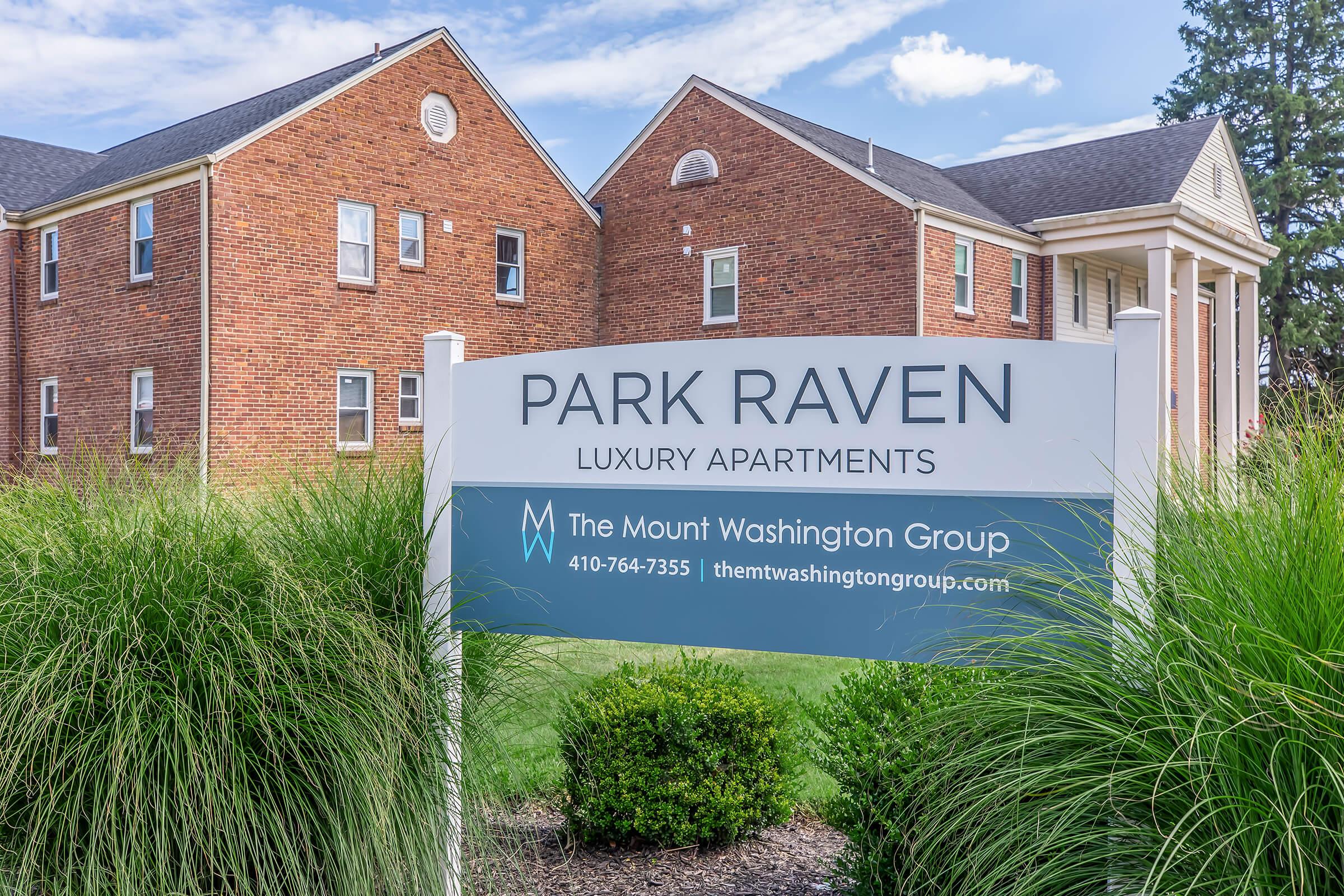 Sign for Park Raven Luxury Apartments, featuring the apartment complex's name prominently displayed in blue. The background includes two brick buildings with a landscaped area of green grass and shrubs. A phone number and website for The Mount Washington Group are also visible on the sign.