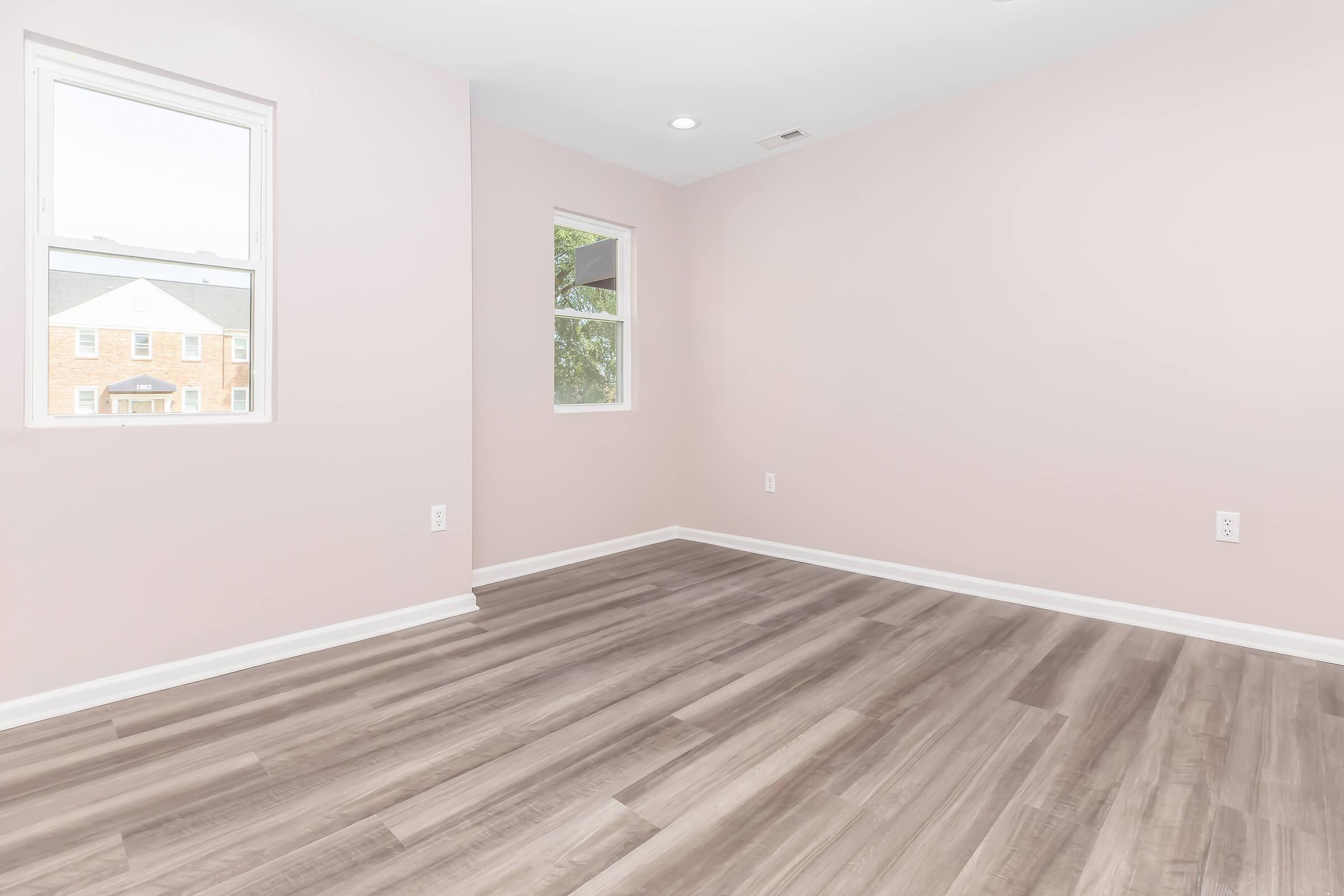 Empty room with pale pink walls and light wooden flooring. Two windows allow natural light to fill the space, framed by white trim. The overall atmosphere is bright and airy, creating a spacious feel. No furniture or decorations present, emphasizing the room's potential for personal design.