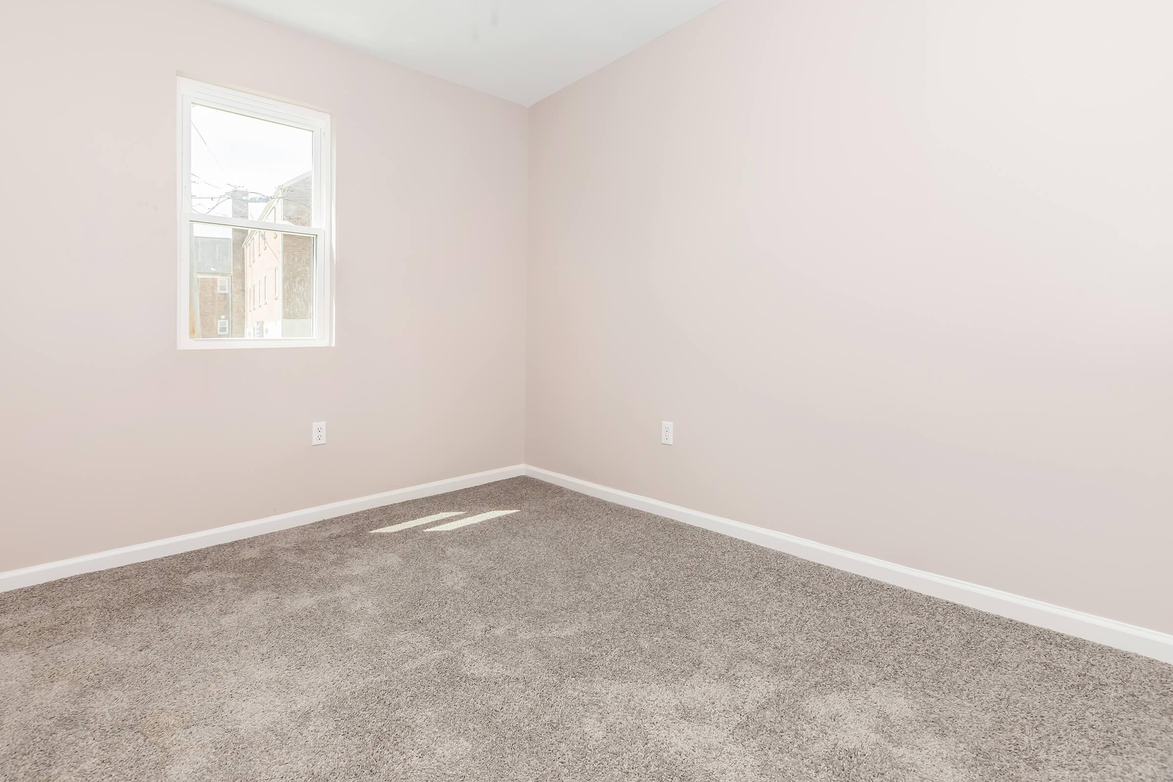 Empty room with light pink walls and a large window allowing natural light in. The floor is covered with beige carpet, and there are no furniture or decorations present, creating a spacious and minimalist feel.