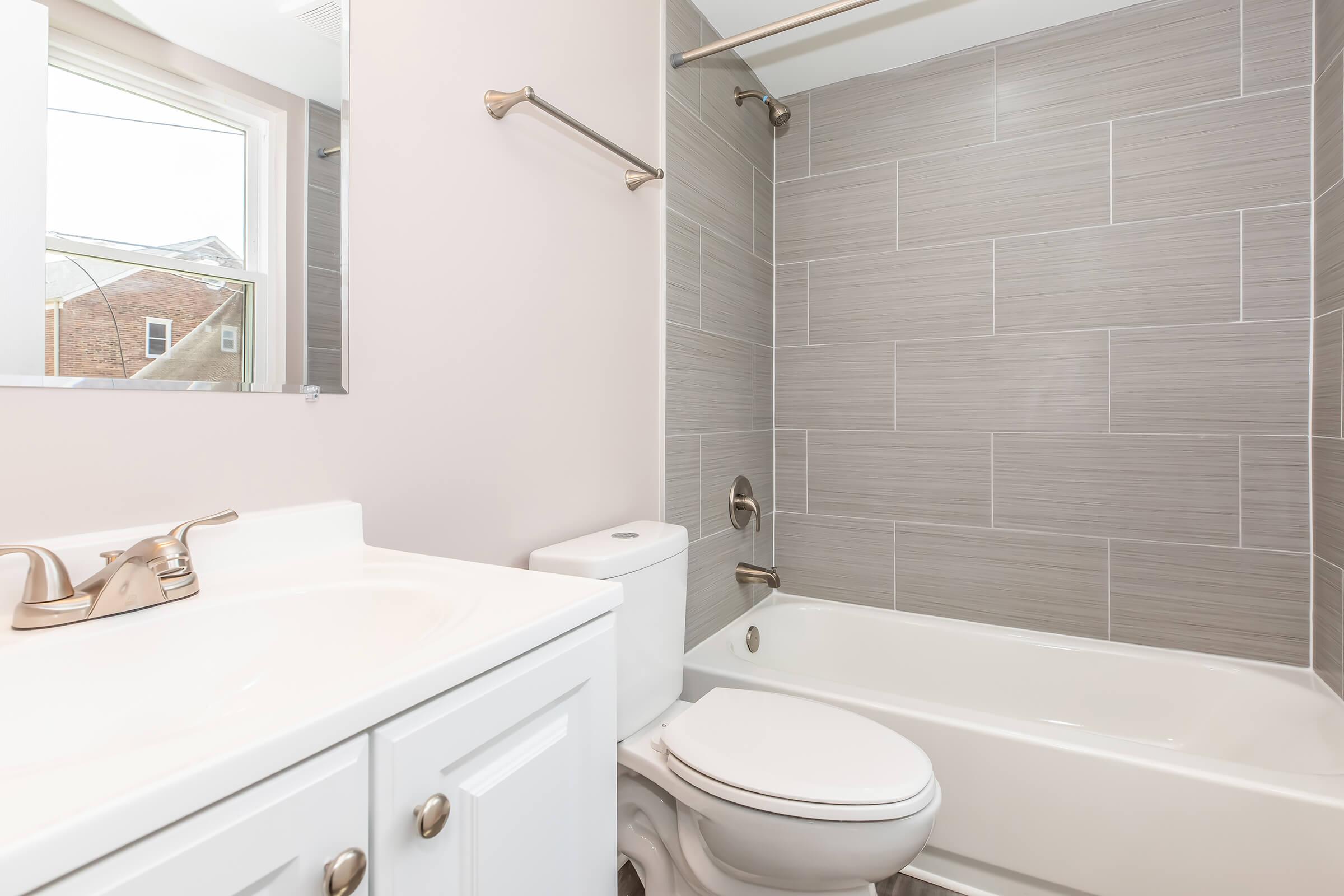 A modern bathroom featuring a white vanity with a sink, a toilet, and a bathtub with a shower. The walls are tiled in light gray, and there is a mirror above the sink. A window allows natural light to enter the space, providing a clean and fresh atmosphere.