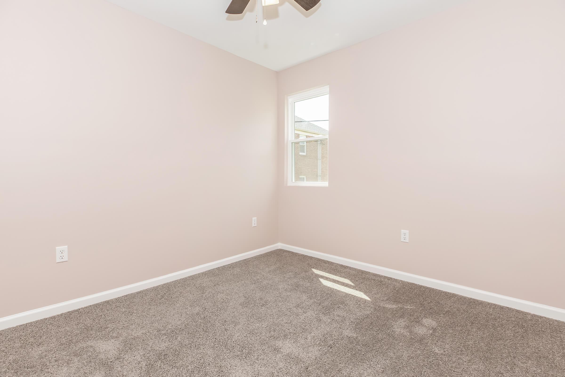 A small room with light pink walls and a light brown carpet. The room has one window letting in natural light and a ceiling fan. There are no furniture pieces, making the space appear empty and minimalistic.