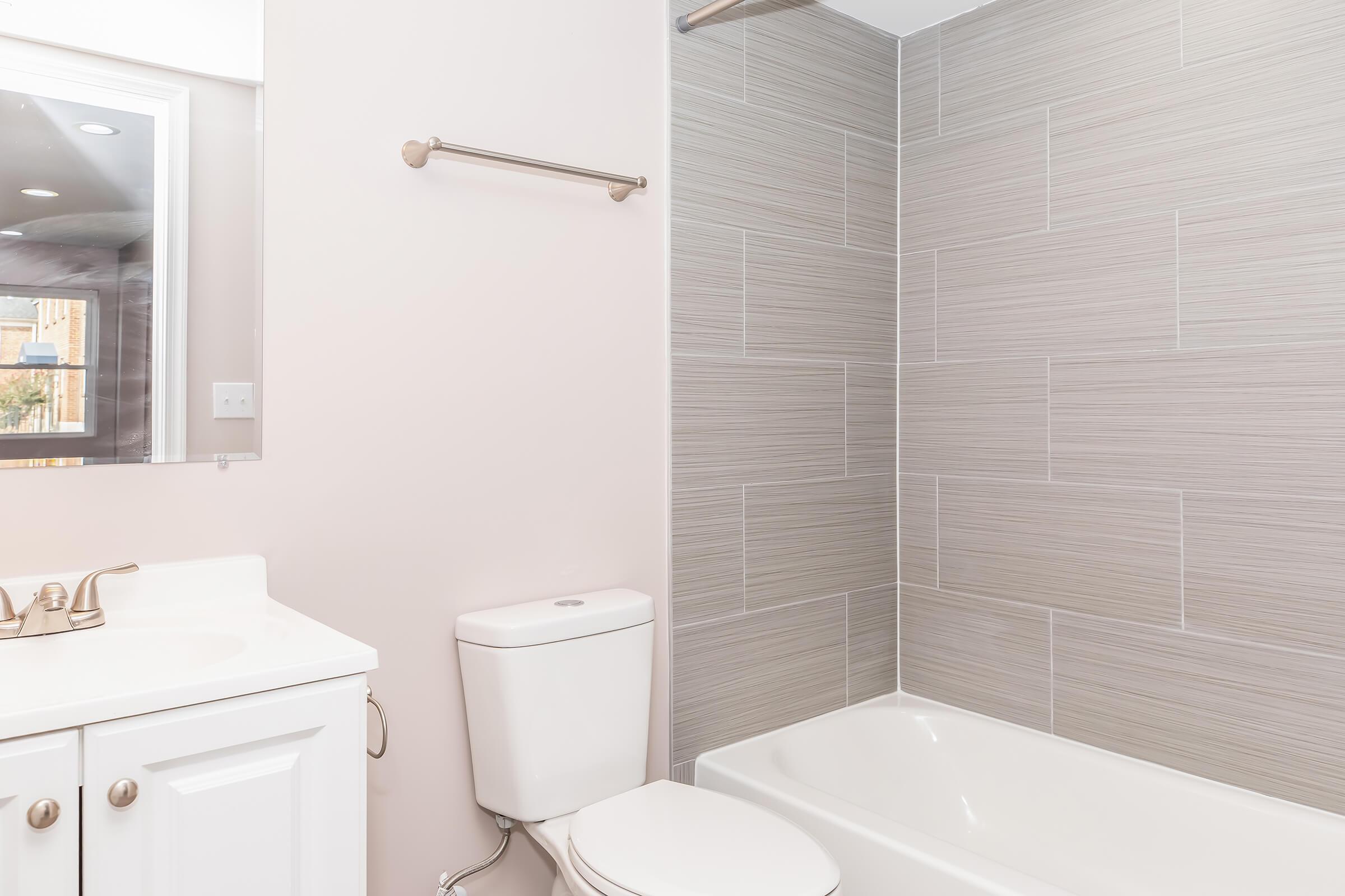 A clean and modern bathroom featuring a white bathtub, a toilet, a sink with white cabinetry, and a mirror. The walls are lined with light gray tiles, creating a fresh and neutral ambiance. Natural light filters in, adding to the spacious feel of the room.