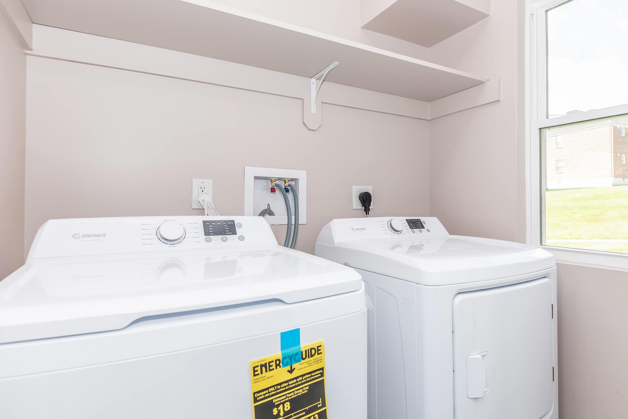A laundry room featuring a white washing machine and a white dryer side by side, with control panels visible. Above, there is a shelf for storage, and a window provides natural light, showing a view outside. The wall is painted in a light color, enhancing the brightness of the space.