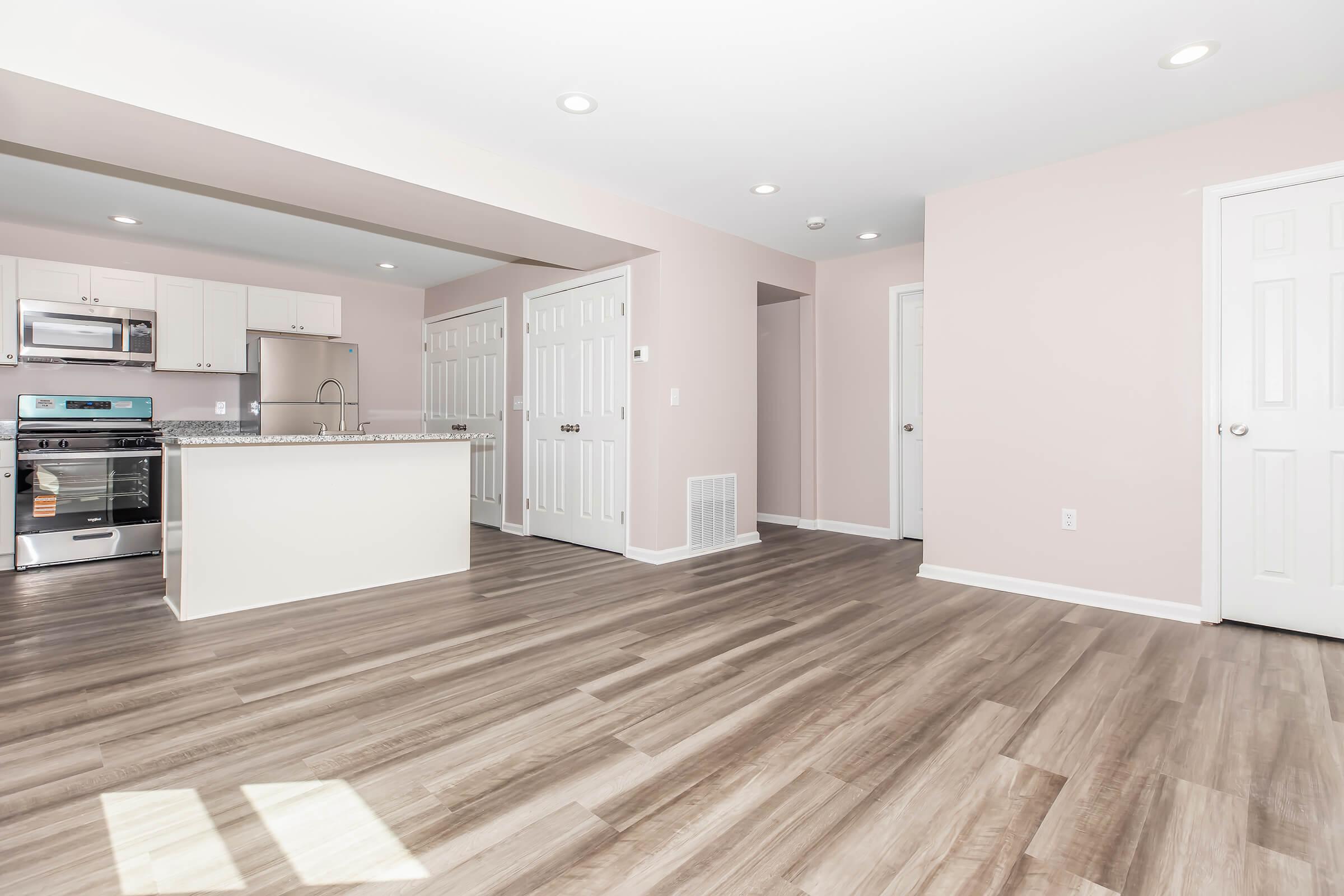 Spacious, modern kitchen and living area with light-colored walls, stainless steel appliances, and a central island. The open layout features laminate flooring and multiple doors leading to storage areas and rooms, creating a bright and inviting atmosphere.