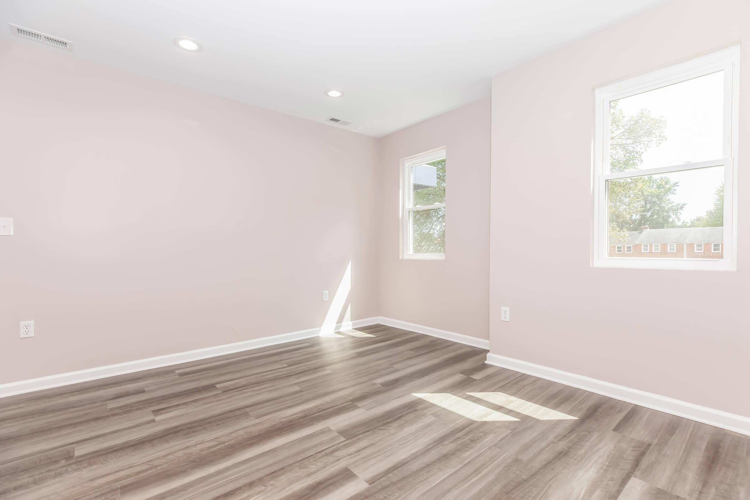 A clean, empty room with light pink walls and large windows allowing natural light. The floor is made of wood-like planks, and sunlight casts shadows on the floor. The space appears fresh and newly renovated, suitable for various uses like an office or bedroom.
