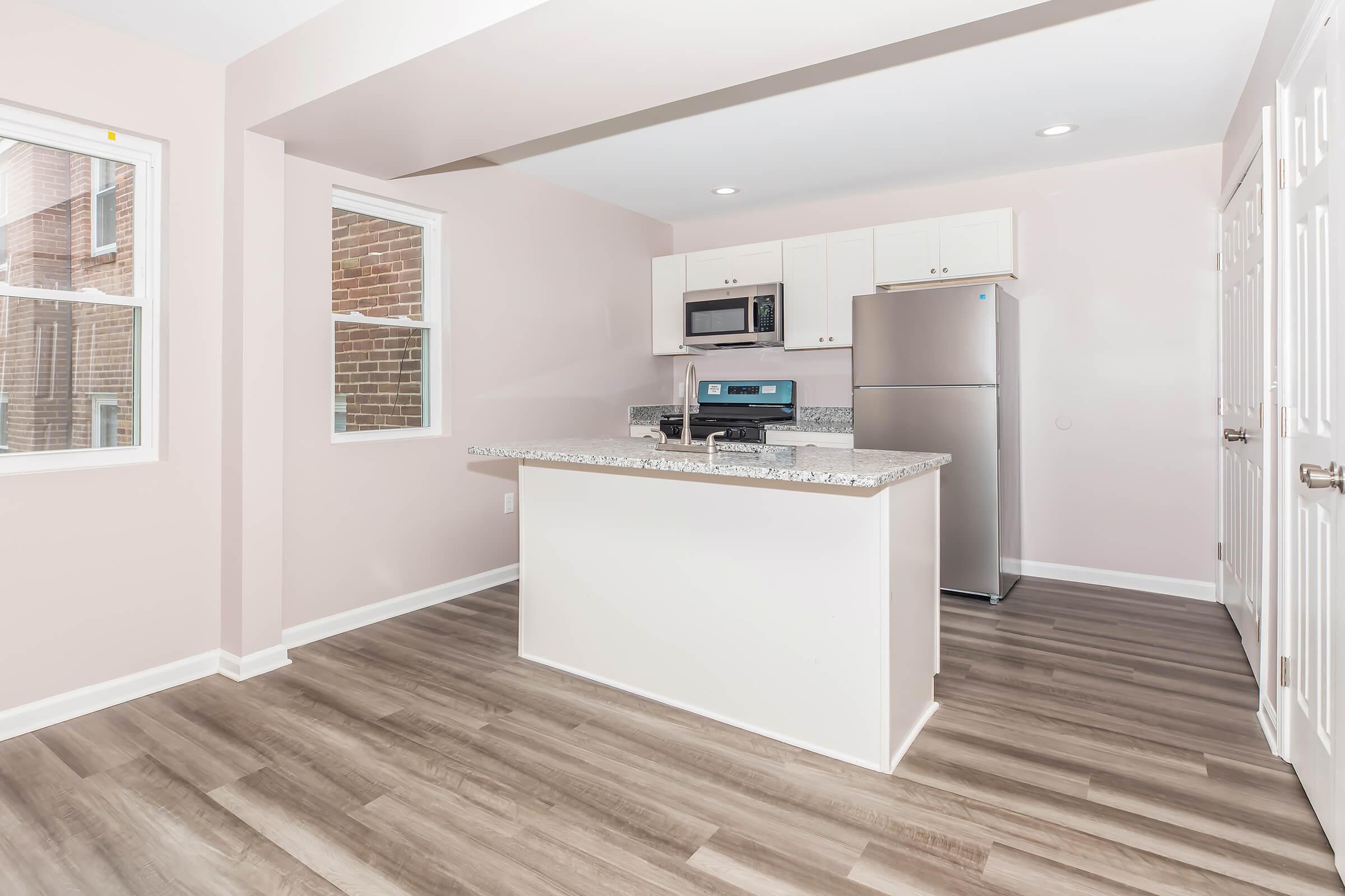 Modern kitchen with light pink walls, featuring a central island with granite countertop, stainless steel appliances including a microwave and refrigerator, and a spacious layout with hardwood-style flooring. Natural light enters through windows on two walls, creating a bright and inviting atmosphere.