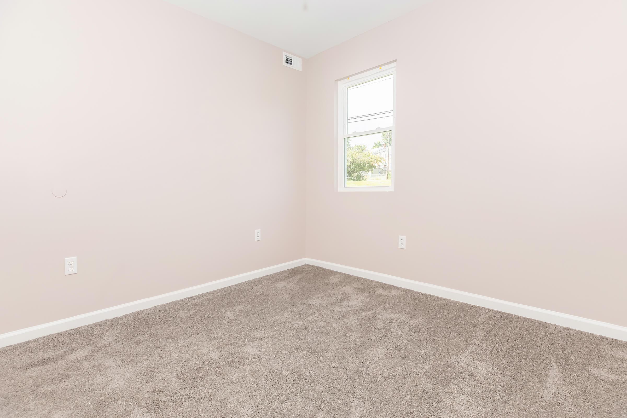 Empty room with light pink walls and gray carpet. One window allows natural light in, illuminating the space. The room has no furniture or decorations, creating a clean and minimalist appearance.