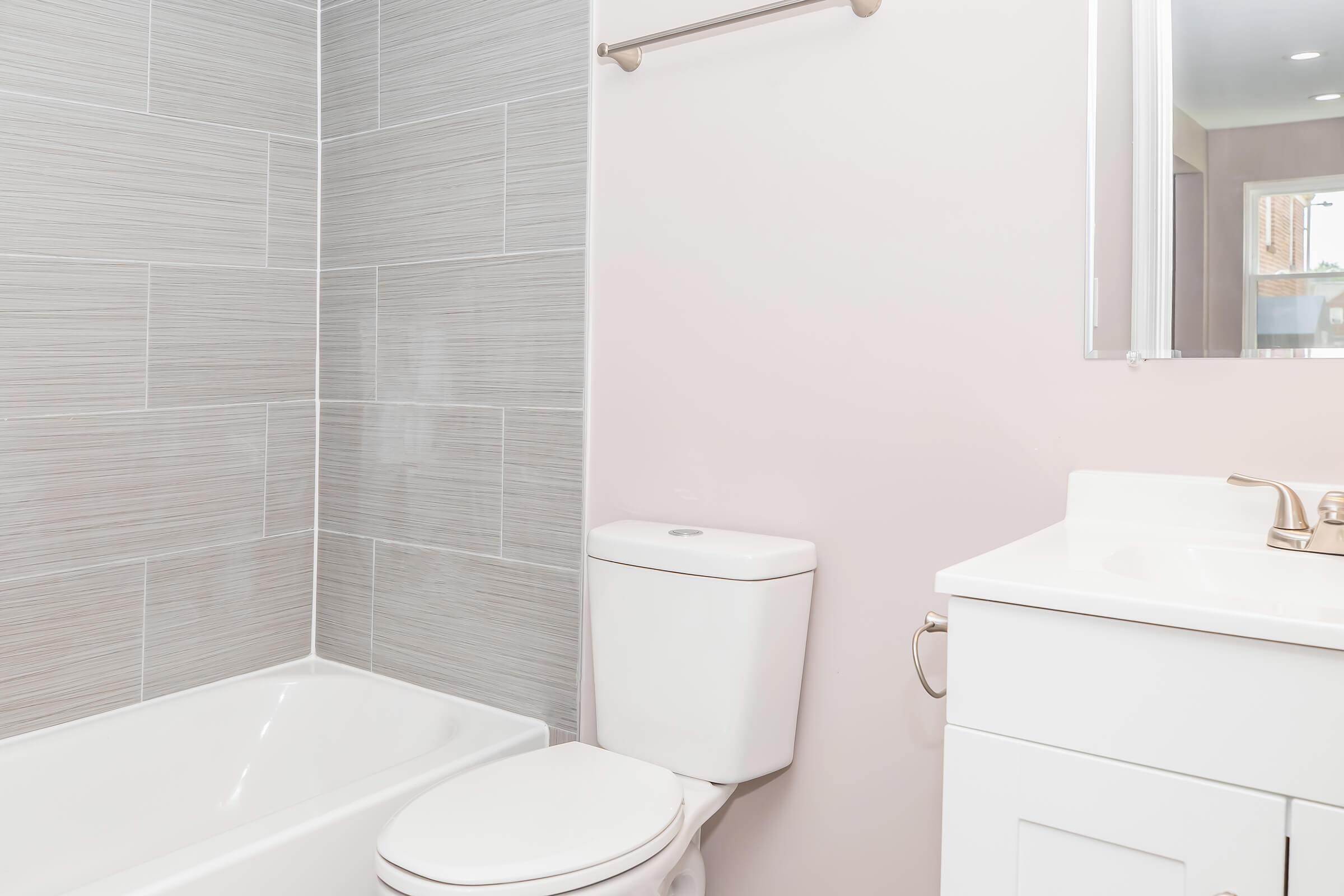 A modern bathroom featuring a white toilet, a bathtub with a gray tiled wall, and a white vanity with a faucet. The walls are light-colored, and there is a mirror above the vanity, creating a clean and contemporary look.