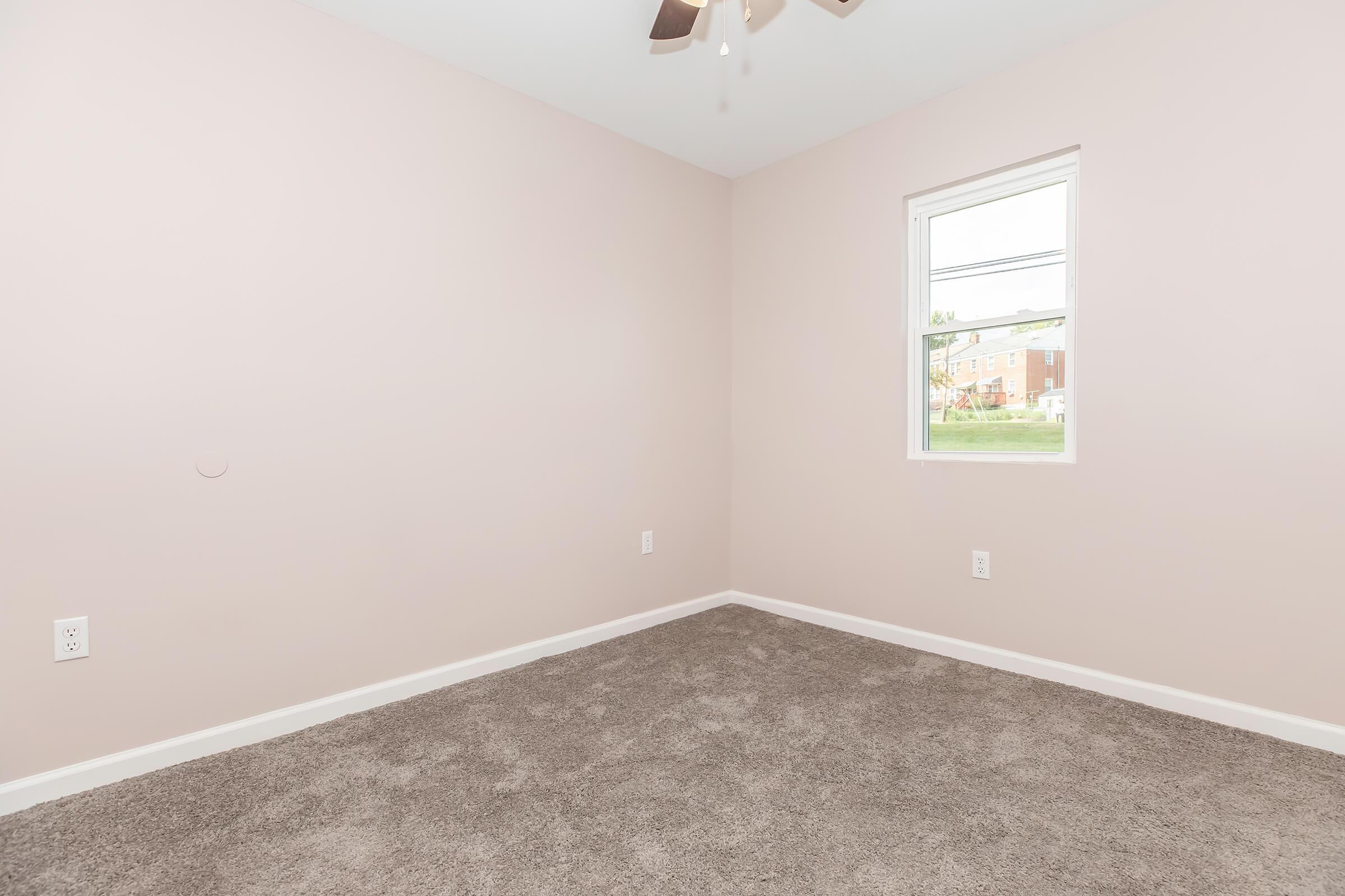 Empty room with light pink walls and a soft carpet. A window on one wall lets in natural light, while a ceiling fan is installed above. The space appears clean and unfurnished, making it suitable for various uses or personal decoration.