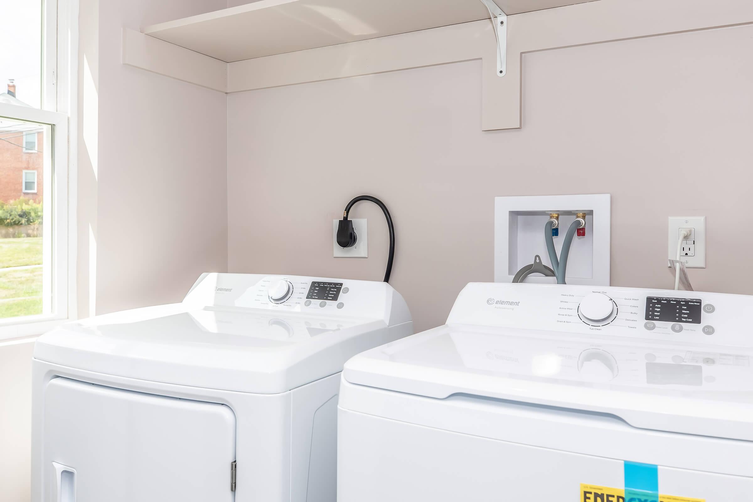A bright laundry room featuring a white washing machine and dryer side by side. The machines have control panels on the front. There's a shelf above them and a window letting in natural light. The wall is painted light pink, creating a clean and modern atmosphere.