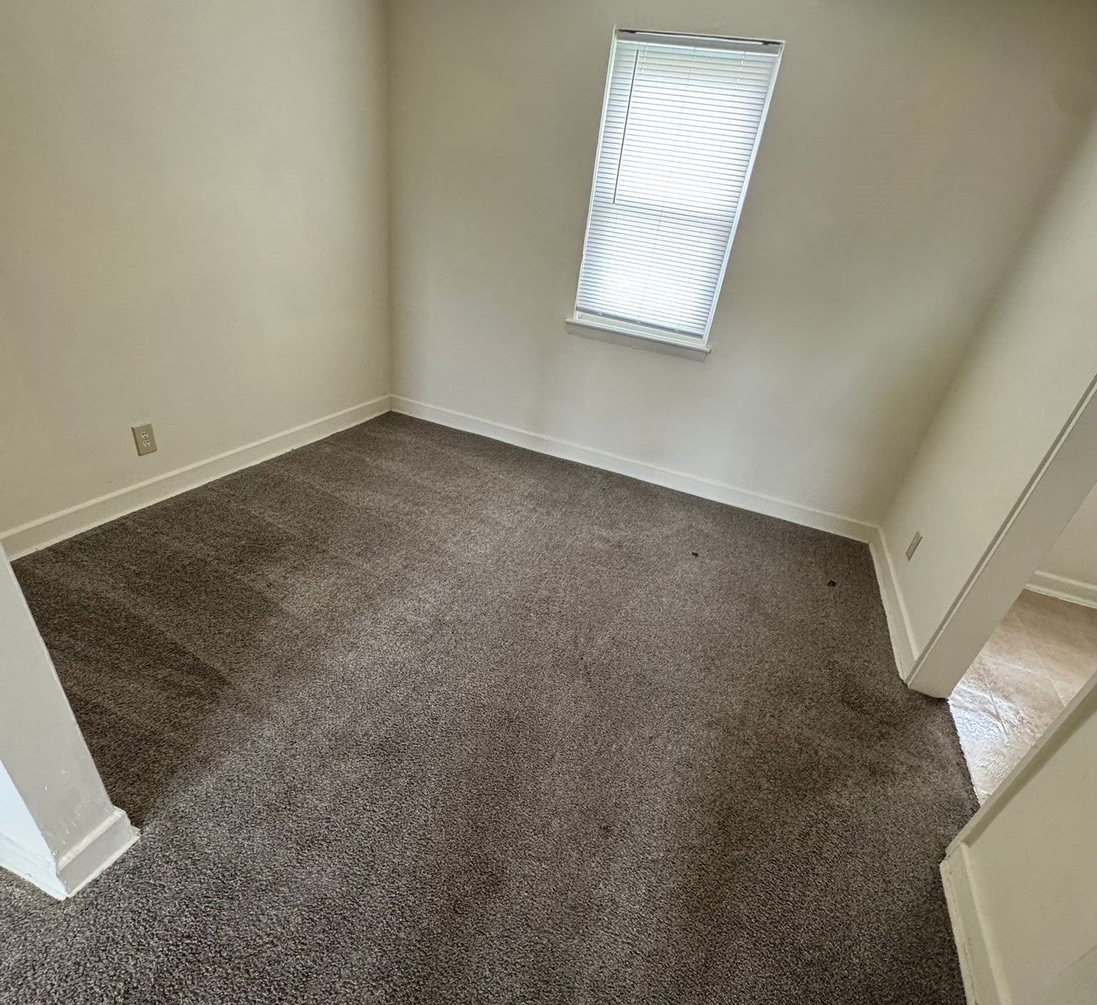 A sparsely furnished room featuring beige carpet and light-colored walls. There is a small window with blinds allowing natural light to enter. The room includes a door leading to another area, giving a sense of space but lacking decorative elements or furniture.
