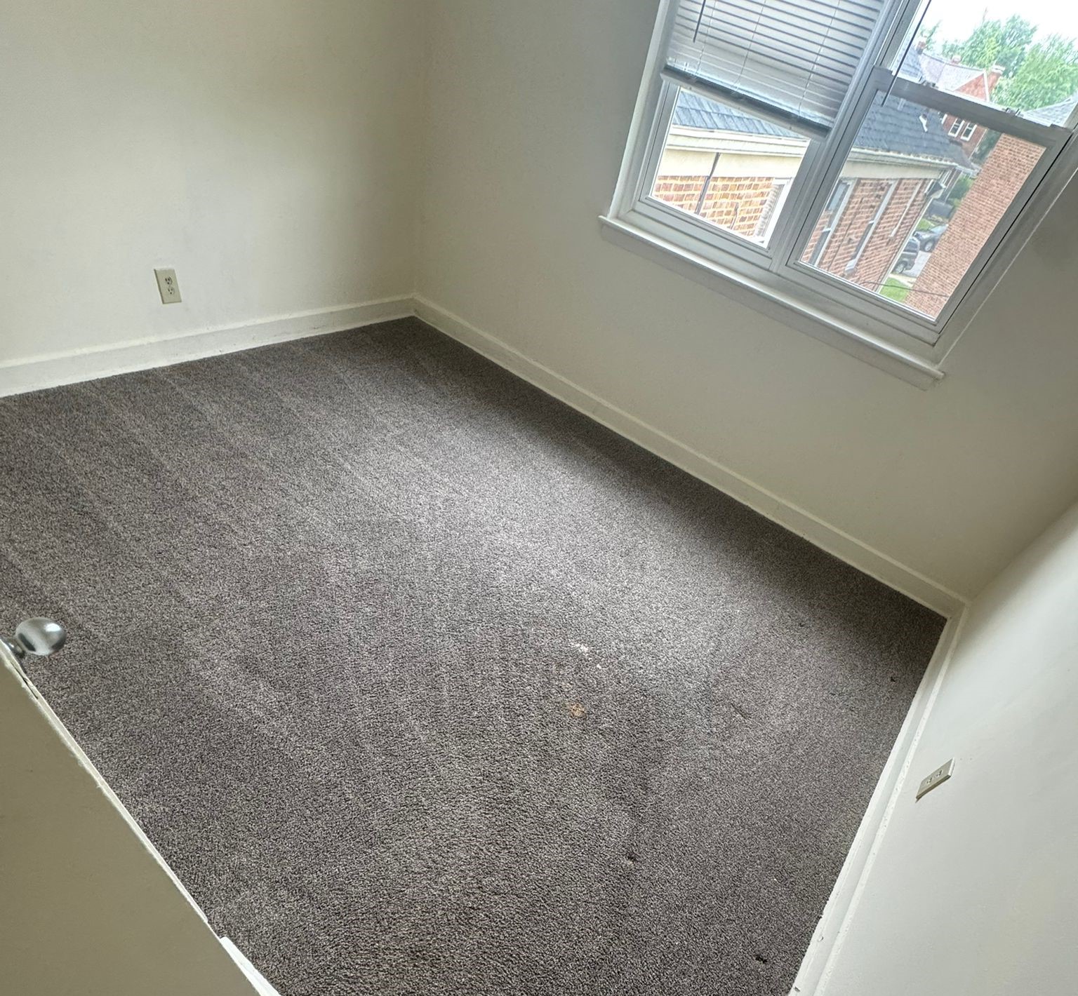 Empty room with light gray carpet, white walls, and a single window allowing natural light. The room has no furniture and features a clean, simple decor, with a few small marks on the carpet. The window offers a view of a neighboring brick building outside.