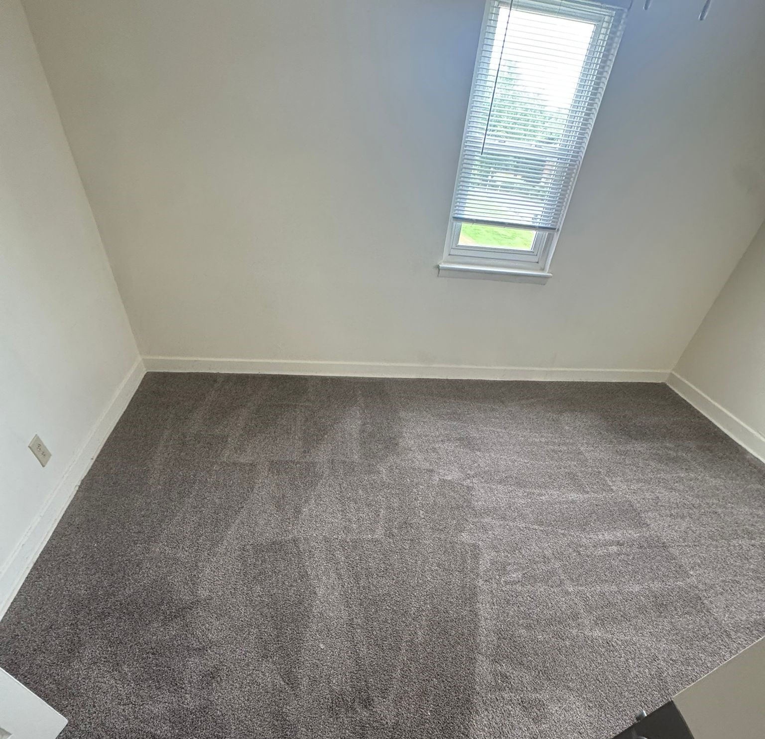 A sparsely furnished room with light gray carpet and white walls. There is a window with blinds allowing natural light in, and no furniture is visible, creating a minimalistic and clean appearance. The room appears ready for use or decoration.