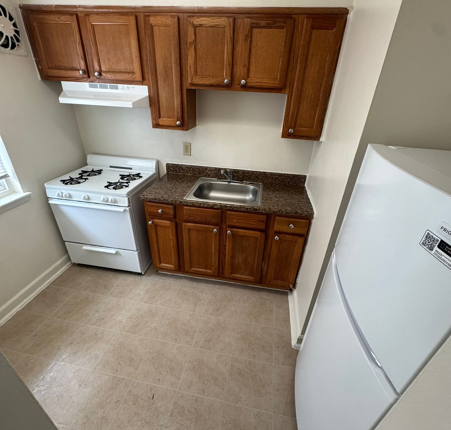 A small kitchen featuring wooden cabinets, a white gas stove, a stainless steel sink, and a white refrigerator. The countertops are dark granite, and there is a window providing natural light. The floor is light-colored vinyl, creating a bright and functional space.