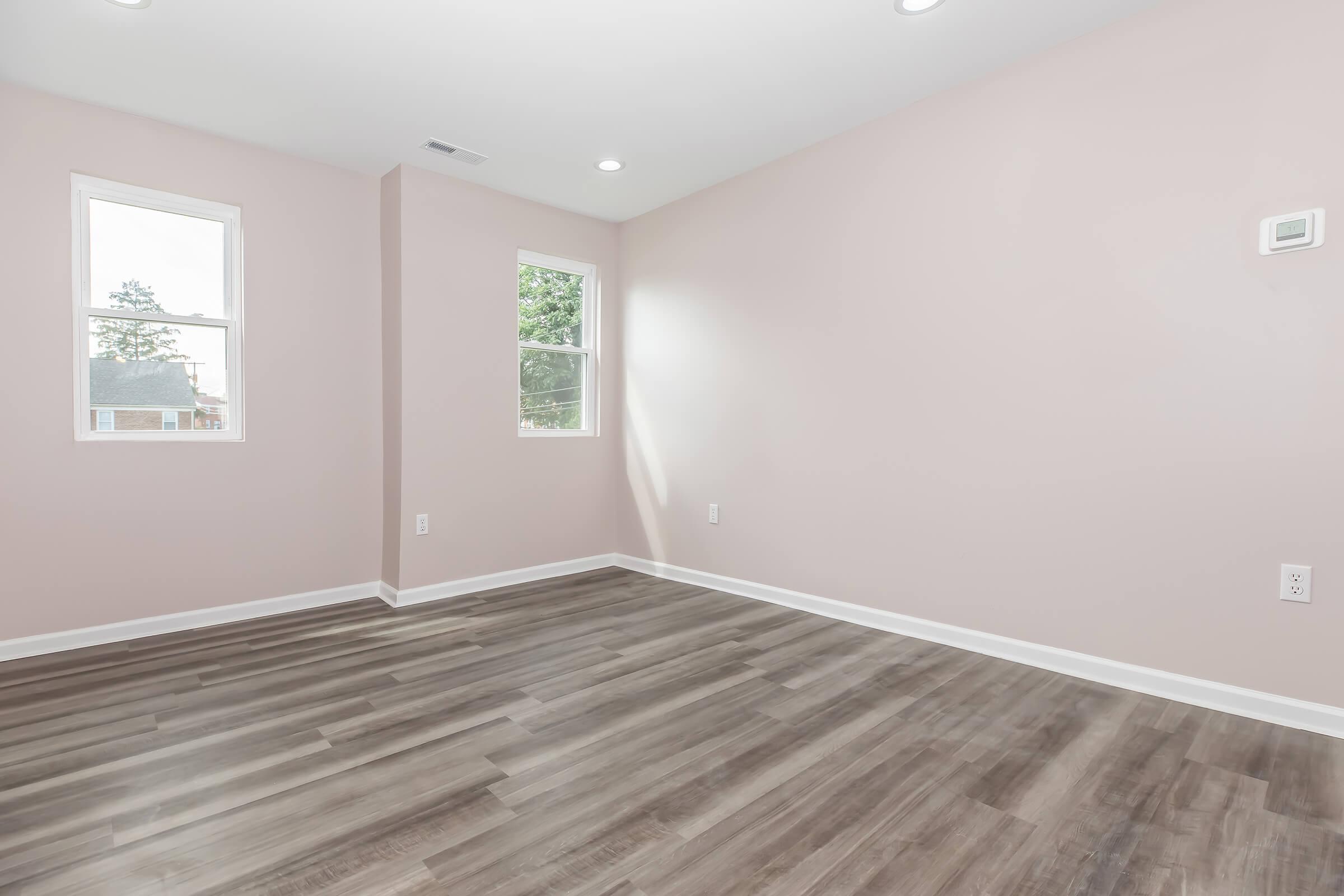 Empty room with light pink walls and two windows allowing natural light. The floor is covered with light brown laminate, providing a clean and modern look. There is a small wall-mounted thermostat on one wall. The space is bright and ready for furnishing.
