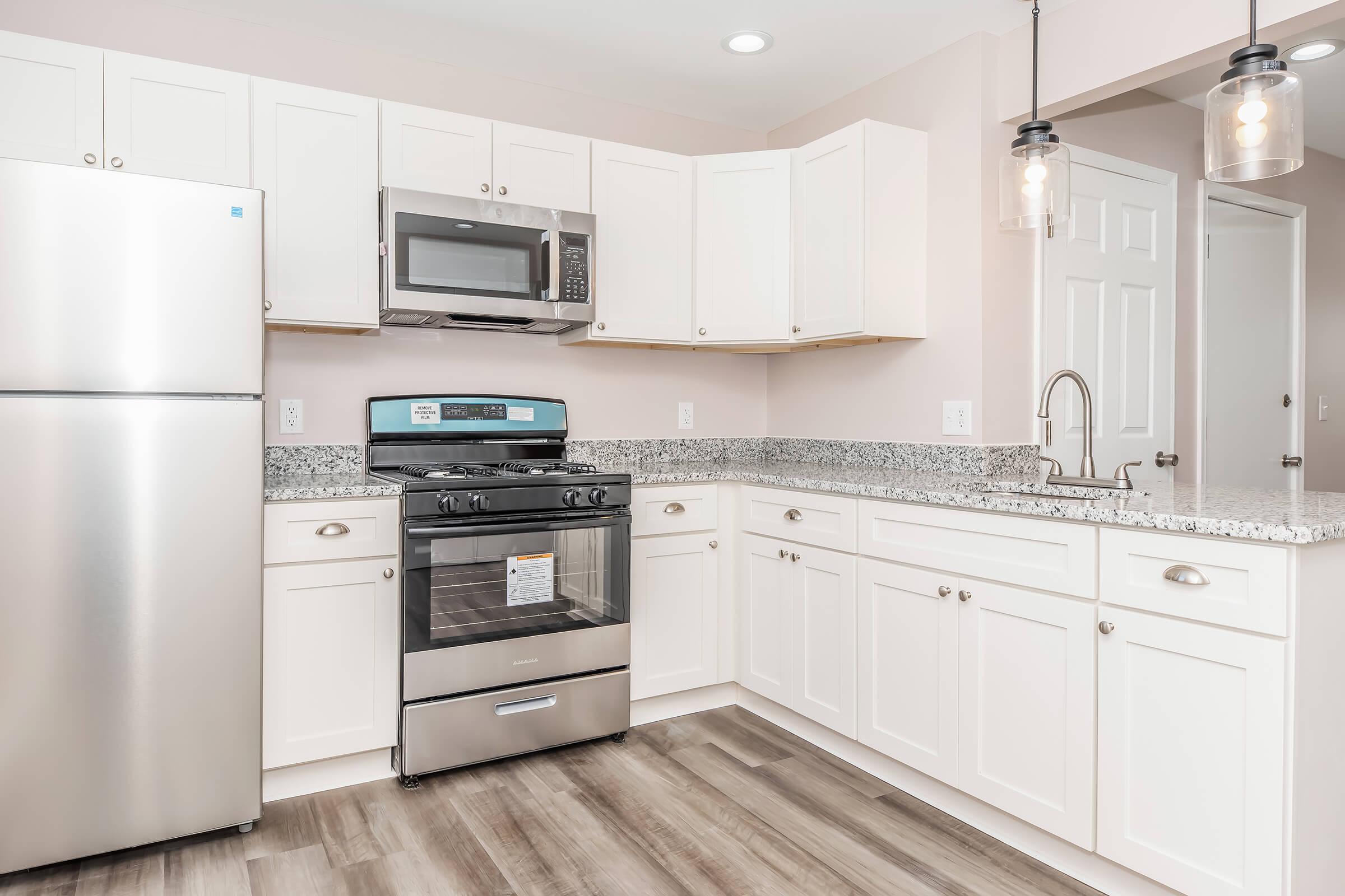 Modern kitchen with white cabinetry, stainless steel refrigerator, microwave, and stove. Granite countertops and pendant lighting add a sleek touch. The floor features a wood-like design, creating a warm atmosphere. Overall, the space appears bright and well-maintained.