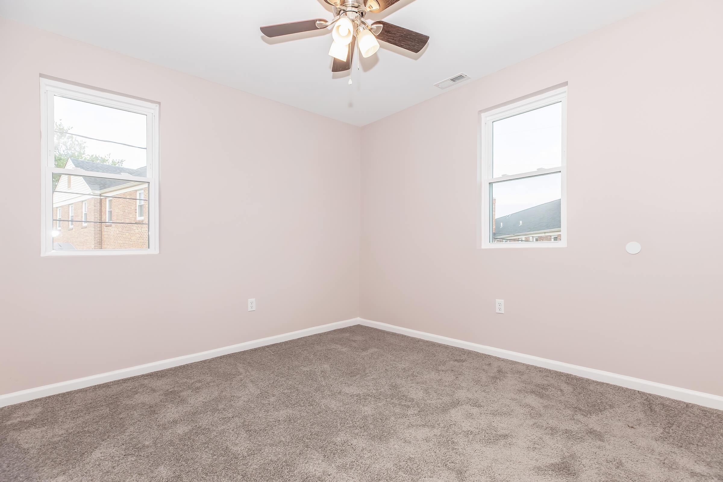 Empty room with light pink walls and gray carpet. Two windows allow natural light, and a ceiling fan with five blades hangs in the center. The room is unfurnished, creating a spacious and inviting atmosphere.