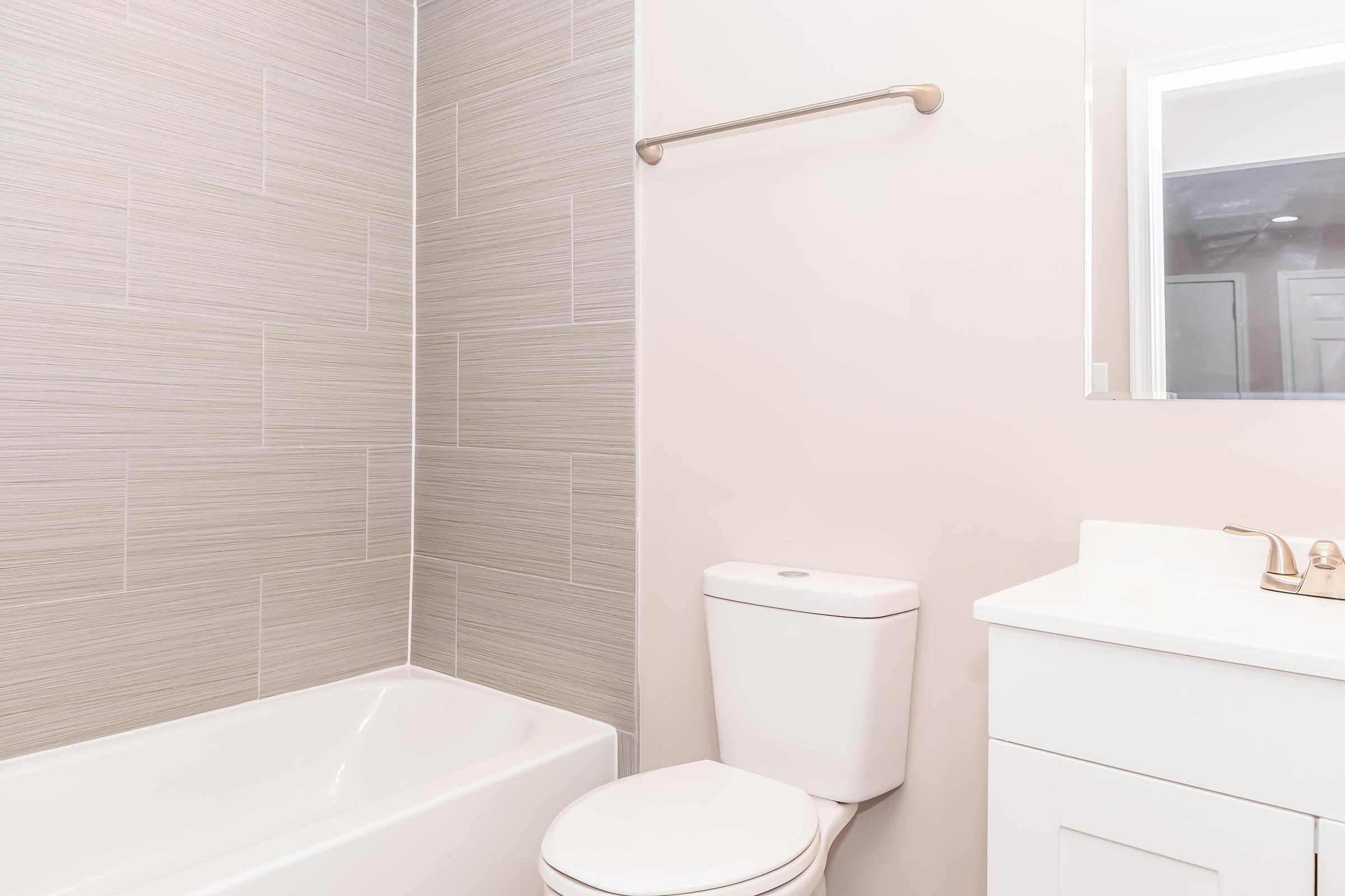 A modern bathroom featuring a bathtub with a tiled wall, a white toilet, and a sleek vanity with a sink and faucet. The walls are painted light-colored, and there is a mirror above the vanity. The overall design is clean and contemporary.