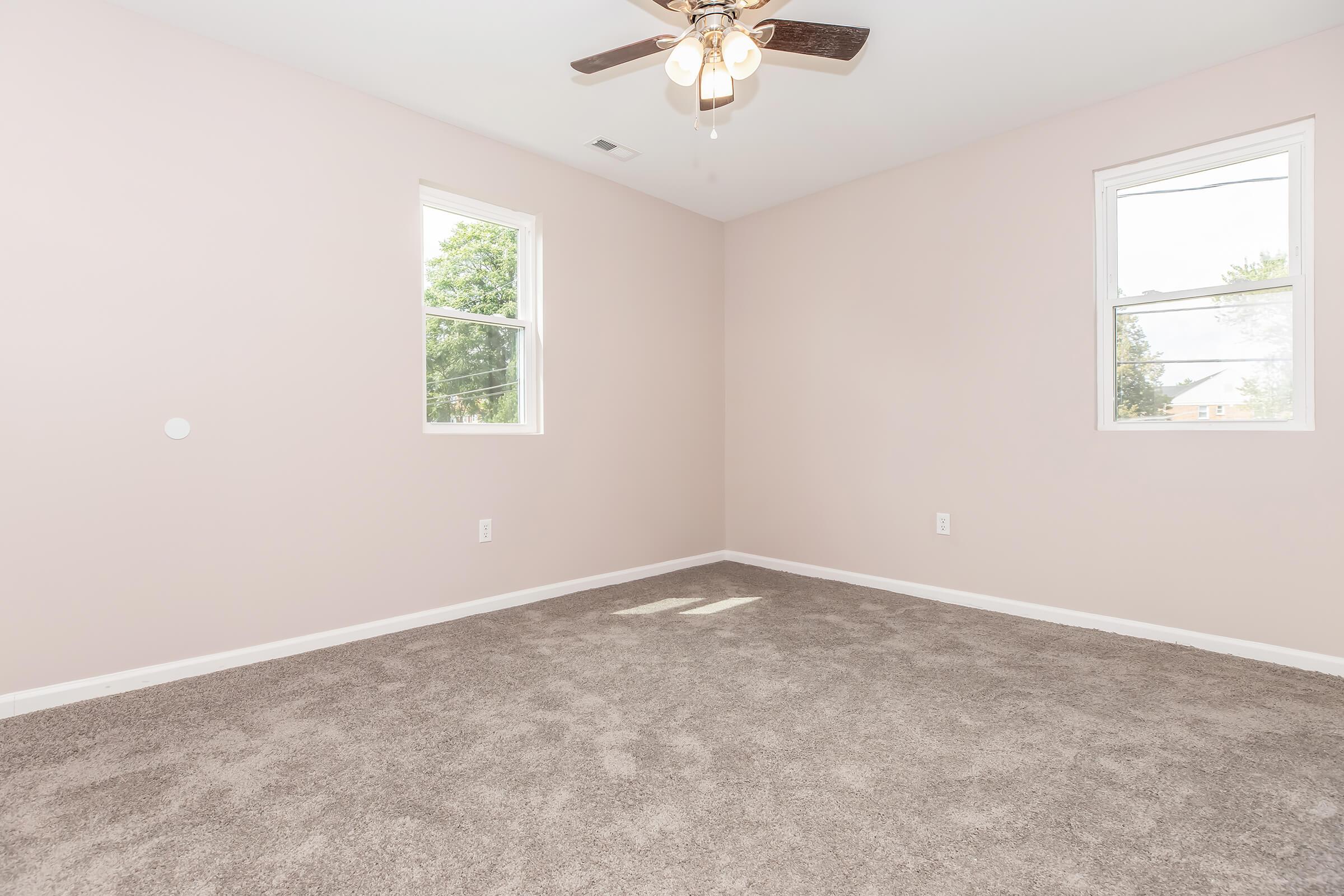 A spacious, empty room with light pink walls, two windows allowing natural light, and a ceiling fan with five blades. The floor is covered in plush beige carpet, creating a cozy atmosphere. The room has a clean and uncluttered appearance, ideal for personal customization.