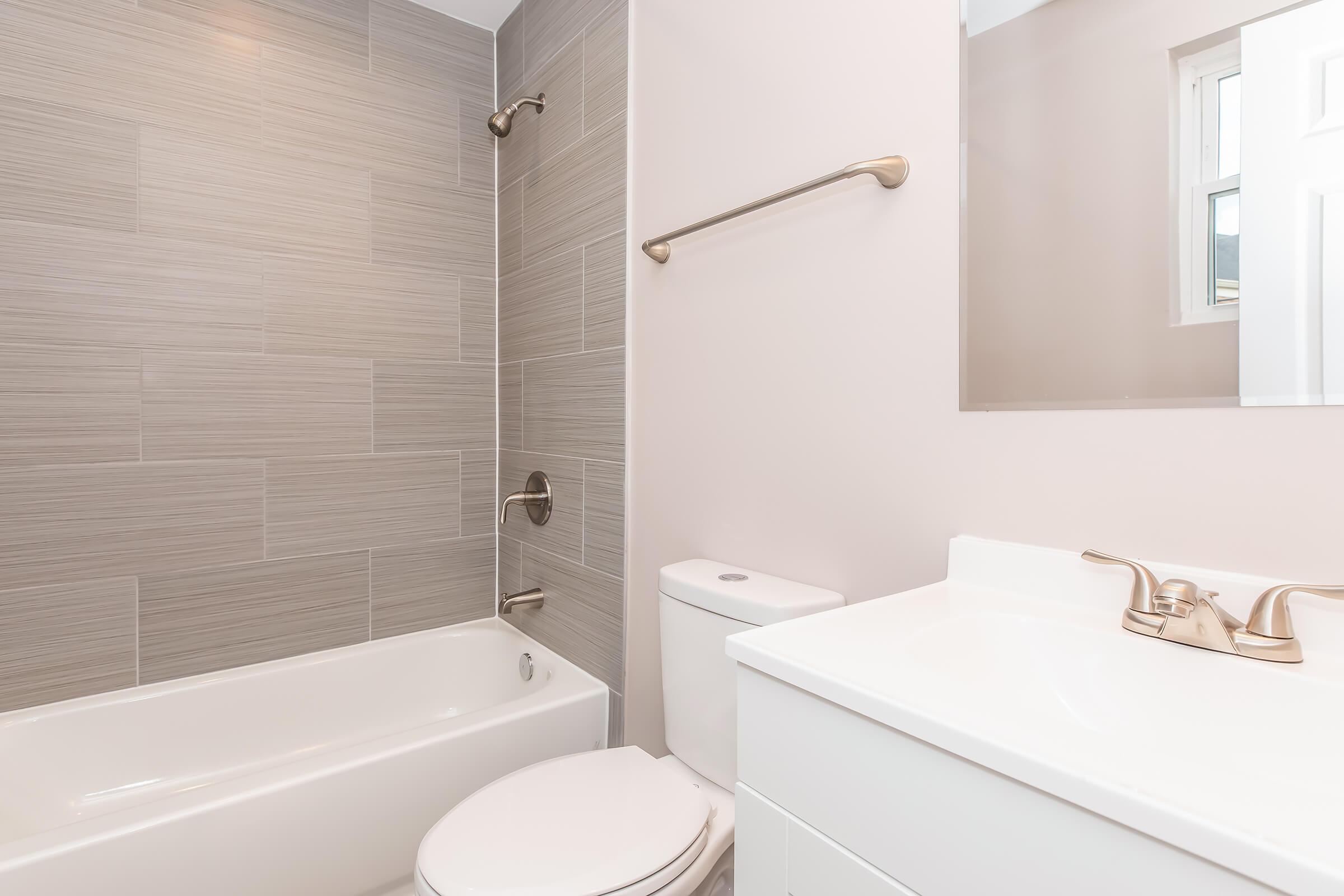 A modern bathroom featuring a bathtub with a shower, sleek tile walls, a toilet, and a white vanity with a sink. The space includes a large mirror and is well-lit by natural light coming from a window. The decor is minimalistic and clean, emphasizing a contemporary design.