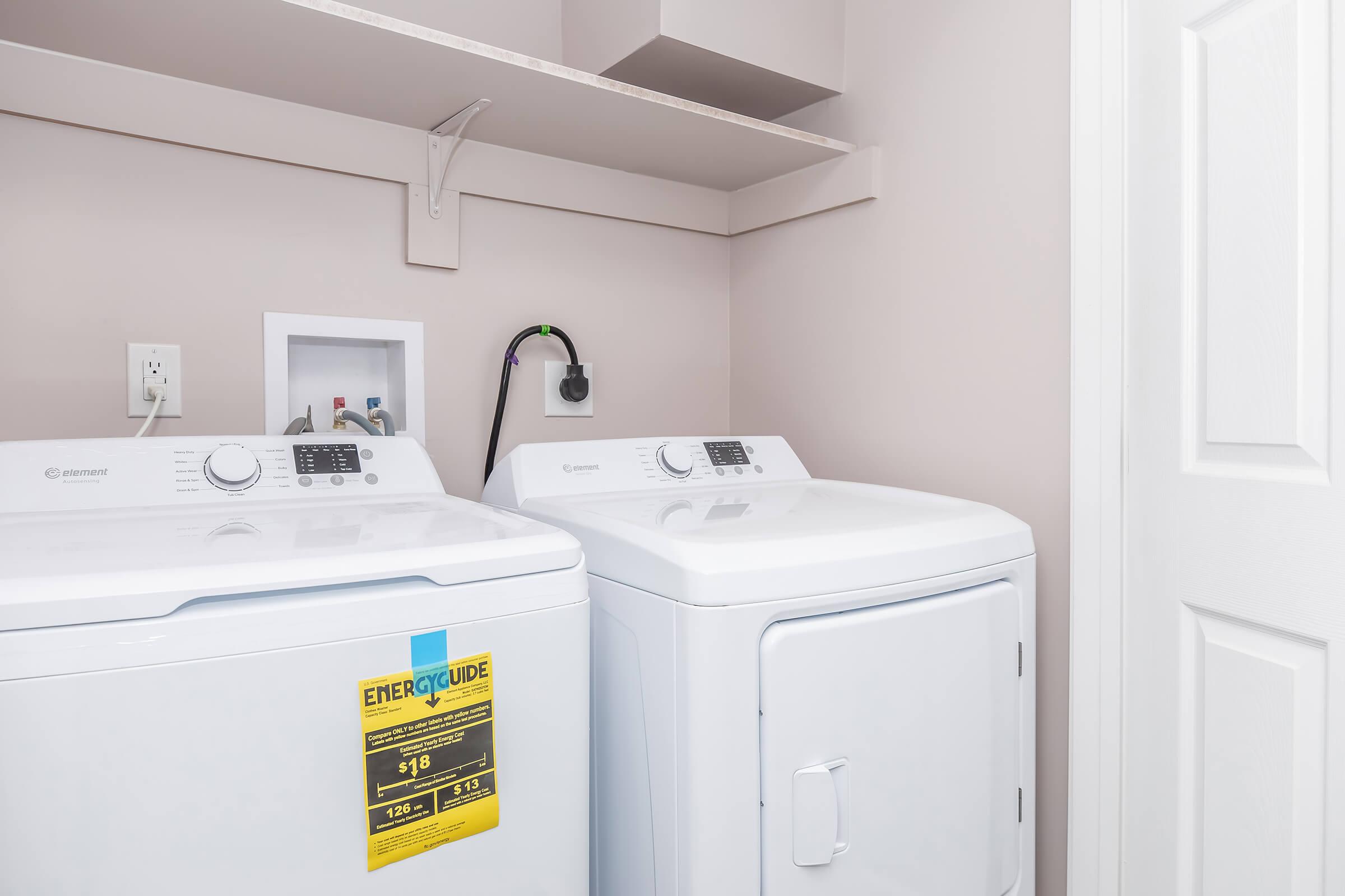 A modern laundry room featuring a washing machine and a dryer side by side. The machines have control panels on top and an energy guide label visible on the dryer. Above them, there's a shelf with minimal items. The walls are painted a soft color, and a door is partially closed in the background.