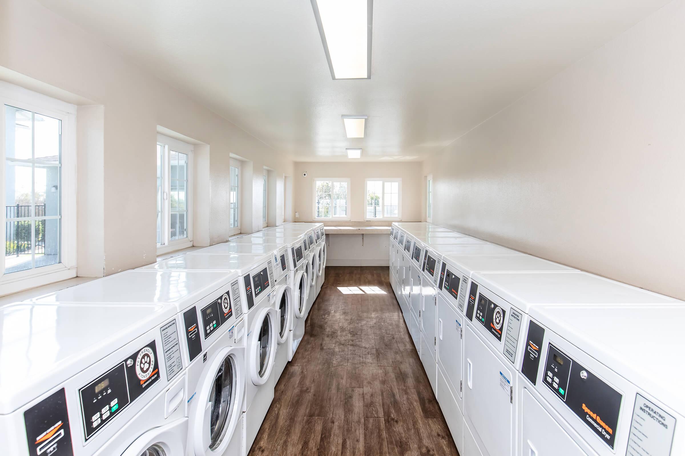 A spacious, well-lit laundry room featuring multiple rows of white washing machines and dryers along the walls, with large windows illuminating the space. The floor is a wood-like finish, creating a clean and modern atmosphere suitable for laundry tasks.
