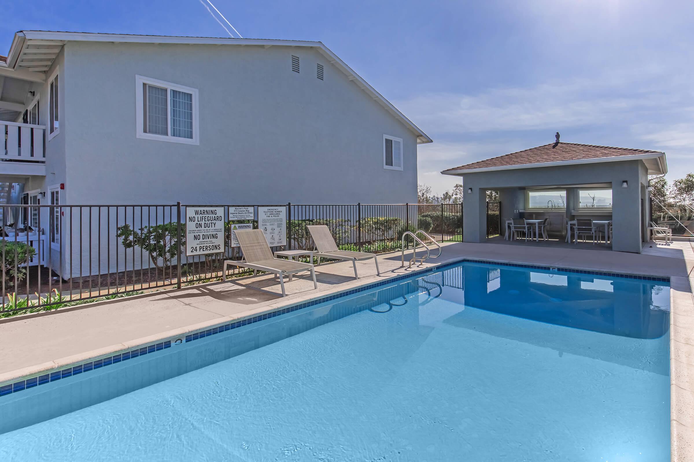 A well-maintained outdoor swimming pool with clear blue water, surrounded by lounge chairs. In the background, there is a light-colored building and a small gazebo or pool house with tables. A fence encloses the pool area, and warning signs are visible nearby. The scene is sunny and inviting.