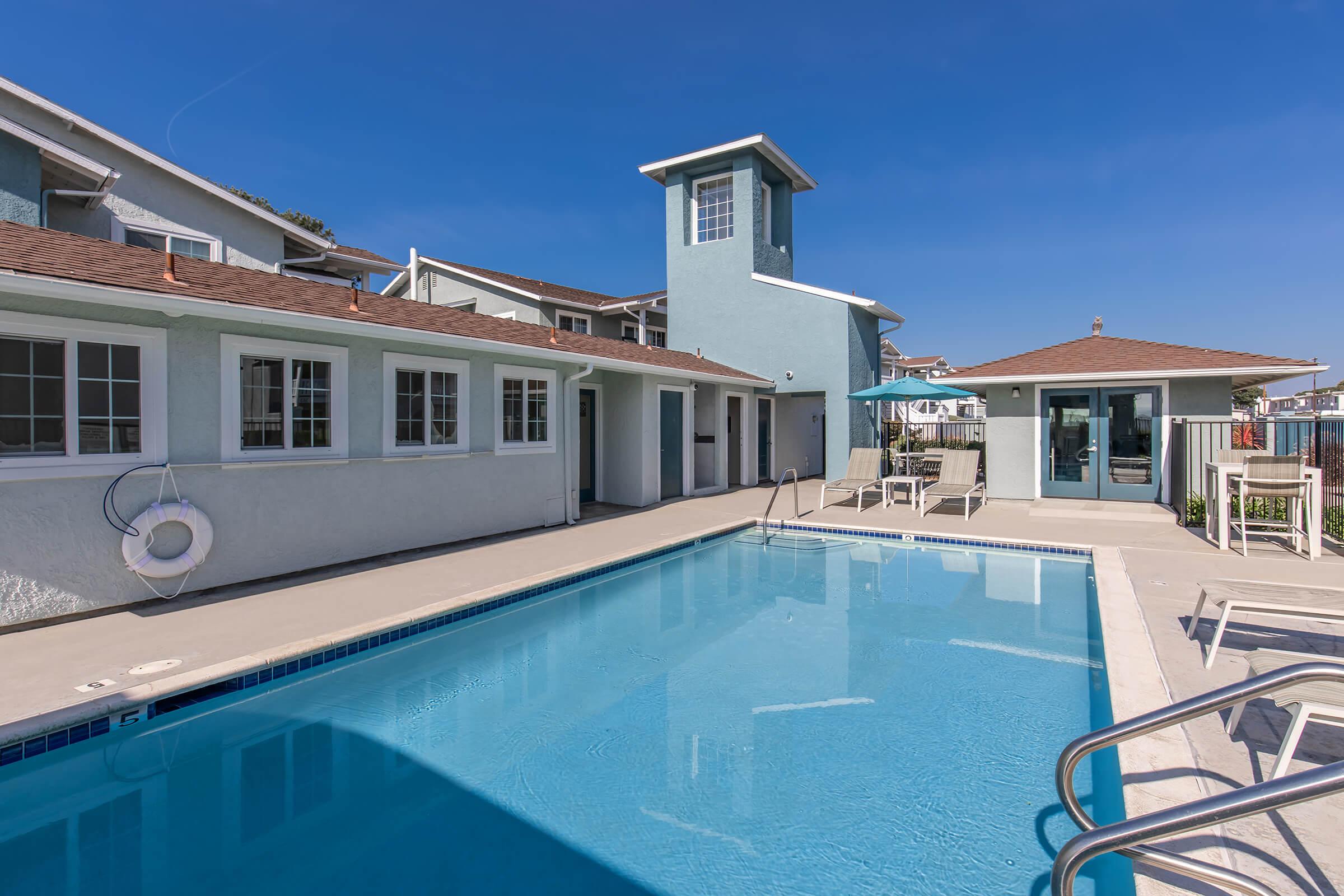 A clear blue swimming pool surrounded by a patio area, with a light blue building and a small gazebo in the background. The sky is bright and sunny, creating a welcoming atmosphere. The scene conveys a relaxing outdoor space ideal for leisure activities.