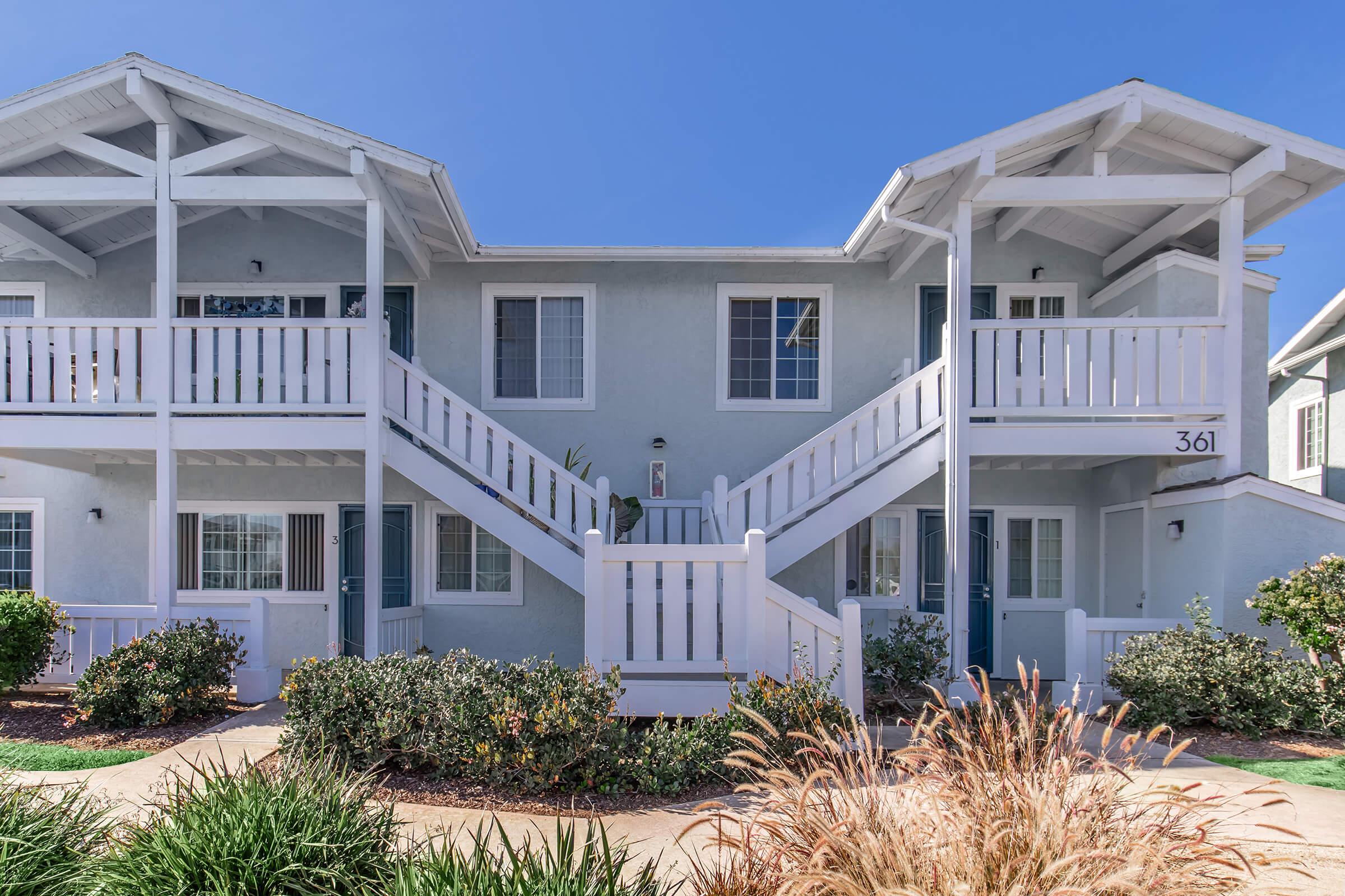 A two-story blue building with white railings and staircases, featuring multiple entrances and windows. The landscape includes neatly trimmed bushes and ornamental grasses, with a clear blue sky in the background. The building has the number "361" displayed prominently.