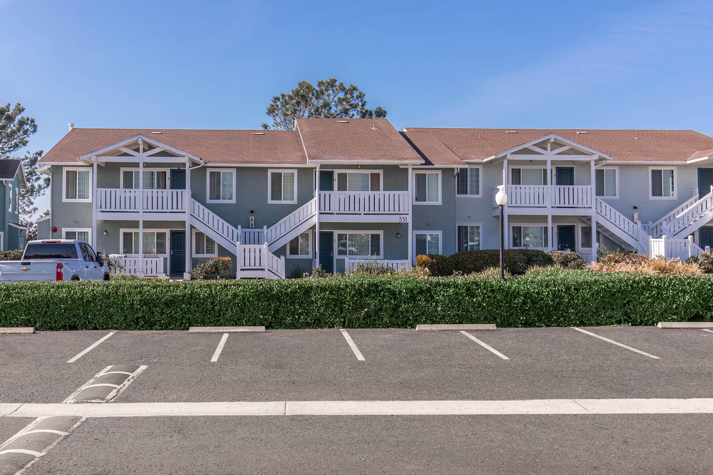 A two-story apartment building featuring a light green exterior with white railings and stairs. It has multiple units visible, surrounded by well-maintained landscaping and parking spaces in front. The sky is clear and blue, contributing to a pleasant atmosphere.