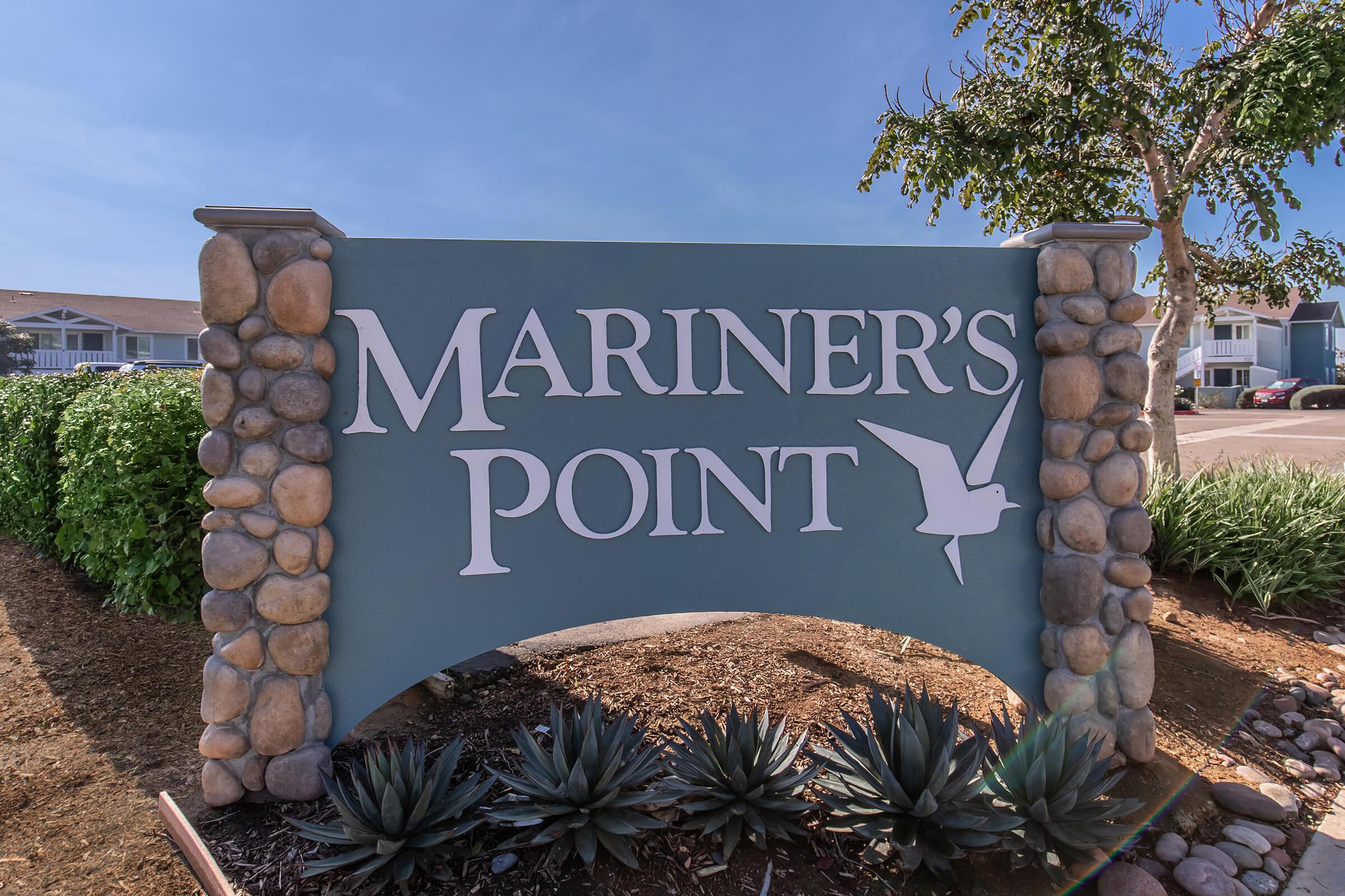 Sign for Mariner's Point with the text prominently displayed. The sign is framed by stone accents and is set against a clear blue sky, with greenery in the background, suggesting a welcoming and serene location.