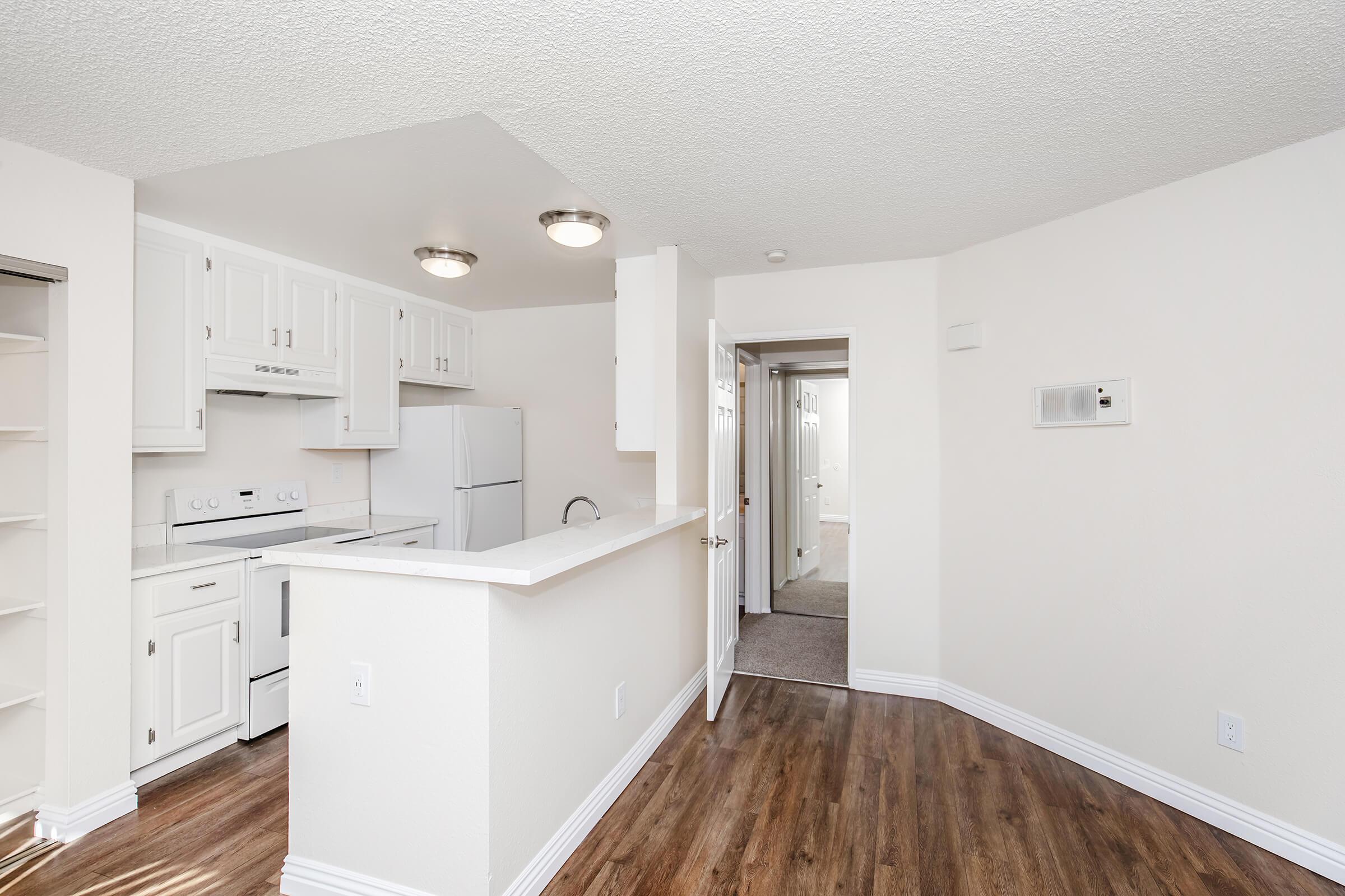 Bright, modern apartment interior featuring an open kitchen with white cabinets, a refrigerator, and a breakfast bar. The space includes a living area with wooden flooring and a hallway leading to other rooms, illuminated by ceiling lights. Clean, neutral tones create a welcoming atmosphere.
