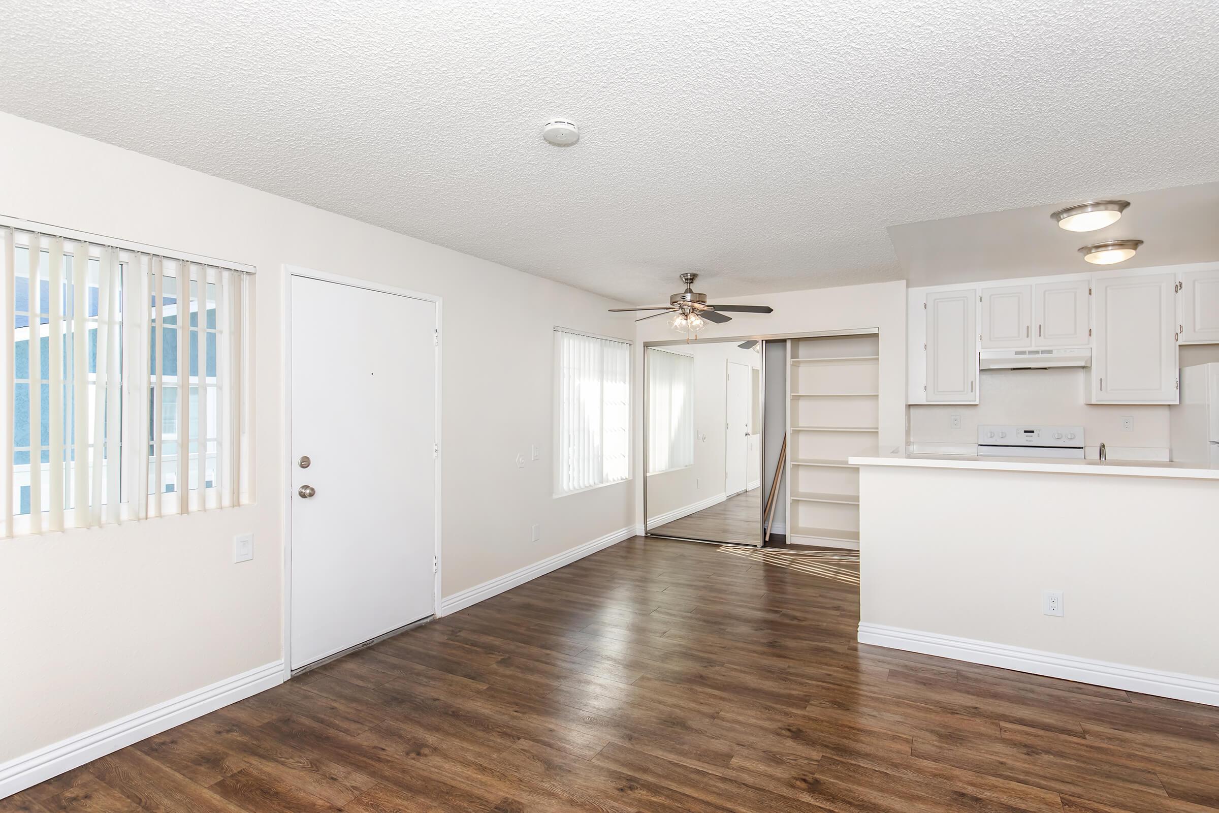 Spacious living area with laminate flooring, featuring a ceiling fan and a partitioned kitchen space in the background. Large windows provide natural light, and the walls are painted in a light color. A door leads to an exterior area, with an open doorway showing additional room access.
