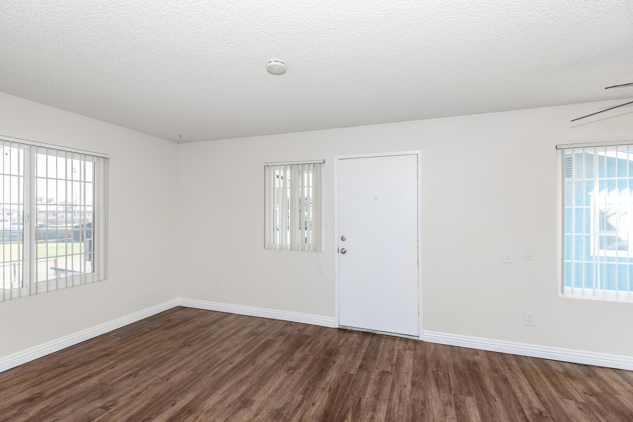 A spacious, empty room featuring light-colored walls, two small windows with blinds, and a white door. The floor is made of wooden planks, and the overall lighting is bright, creating an open and airy atmosphere. The room is ready for furnishing and personalization.