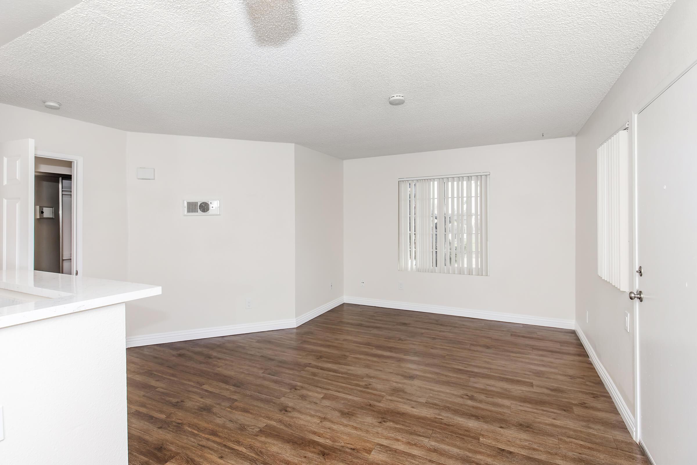 A spacious, empty living room with light-colored walls and a textured ceiling. The floor features wood-like laminate. A small air conditioning unit is mounted on one wall, and there are vertical blinds on a window. The room is well-lit with natural light, creating a bright and inviting atmosphere.