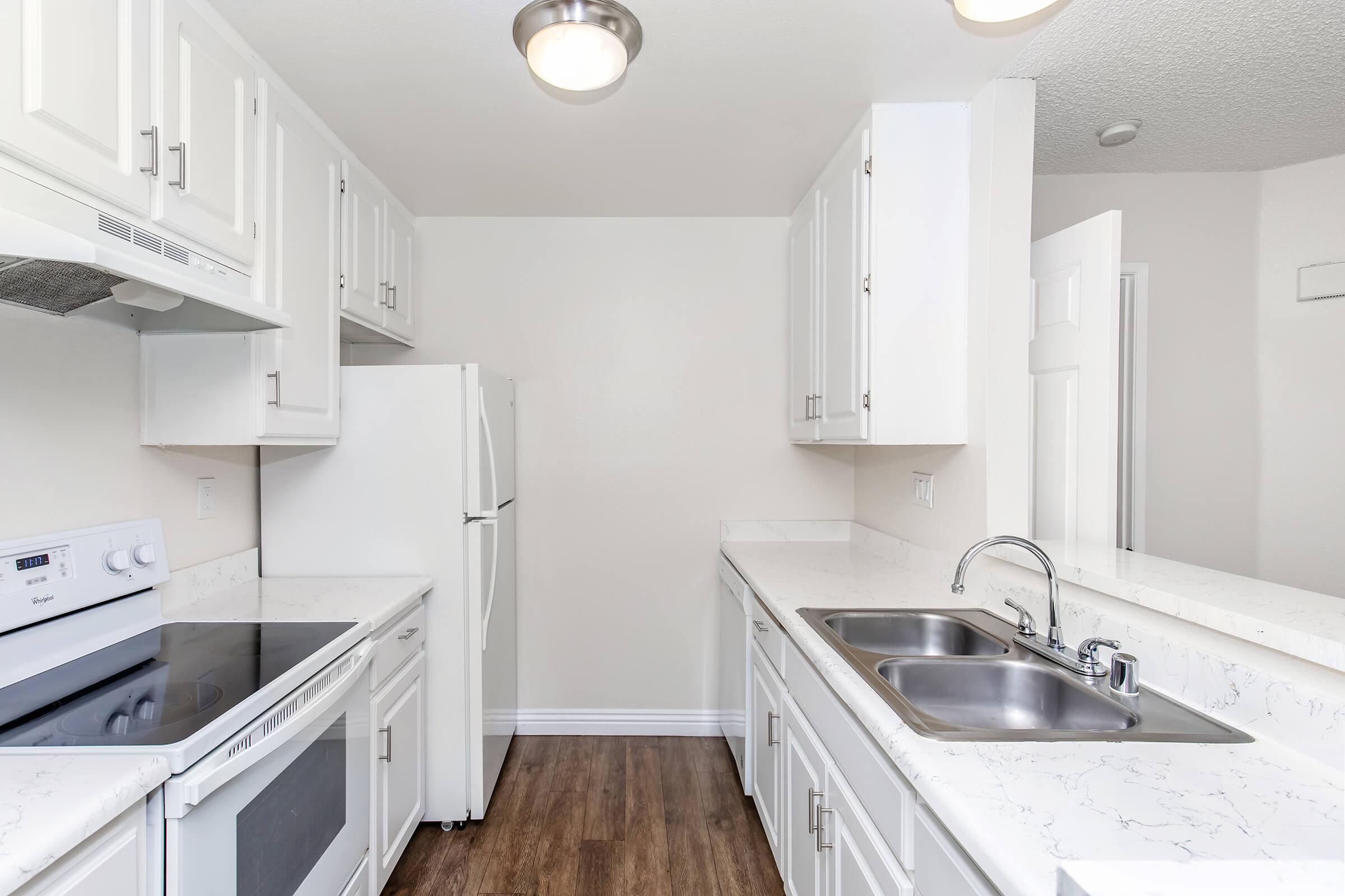 Modern kitchen featuring white cabinetry, a stainless steel sink with a double basin, and a white refrigerator. Countertops are light-colored with a marble effect. The kitchen includes an electric stove and an overhead microwave, all set in a bright, clean environment with wood-like flooring.