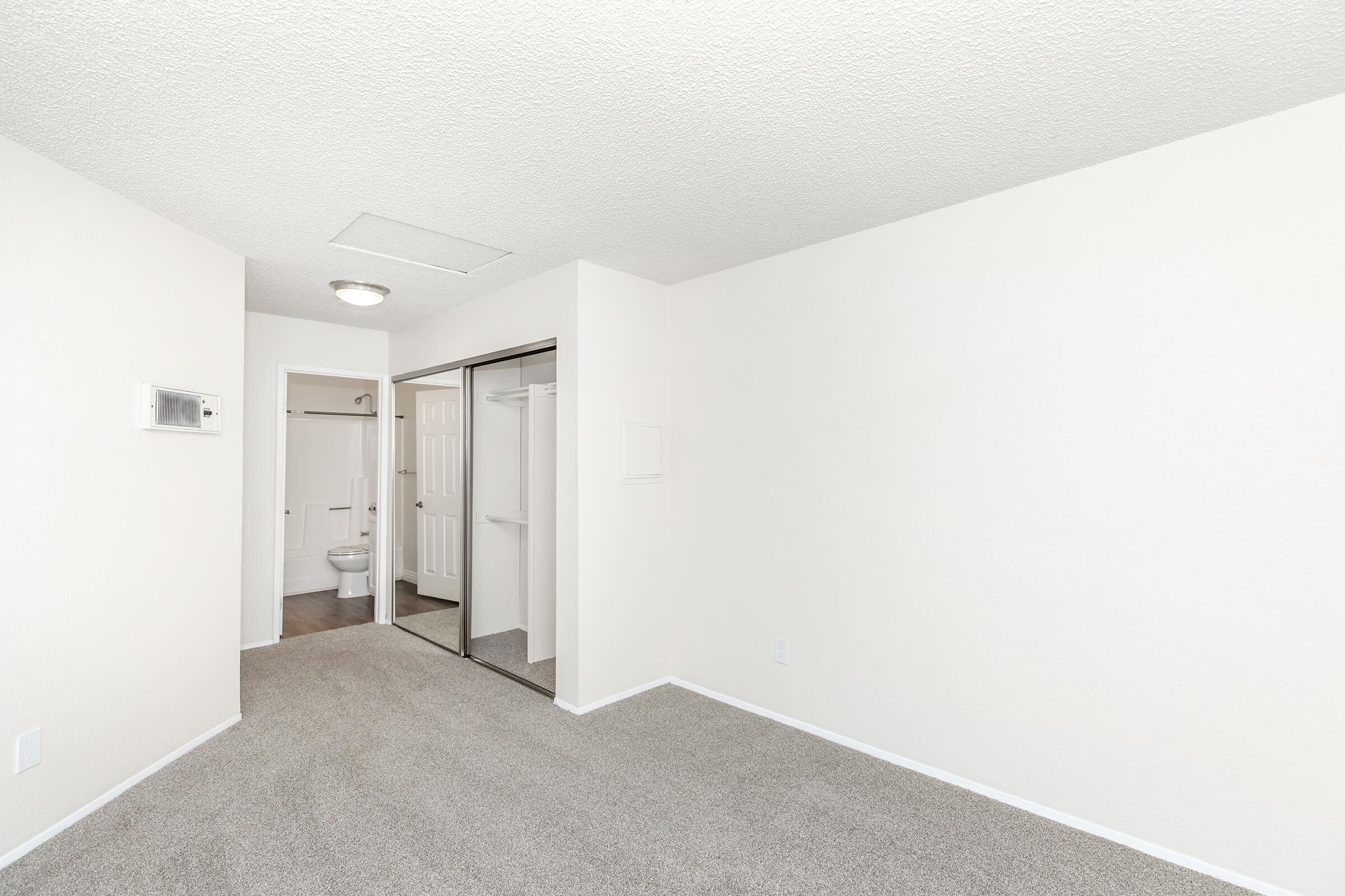 Empty room with beige carpet and white walls. A mirrored sliding door leads to a small closet, and a bathroom is visible in the background. The ceiling features a light fixture, and there's a small air conditioning unit on the wall. The overall space is bright and clean.
