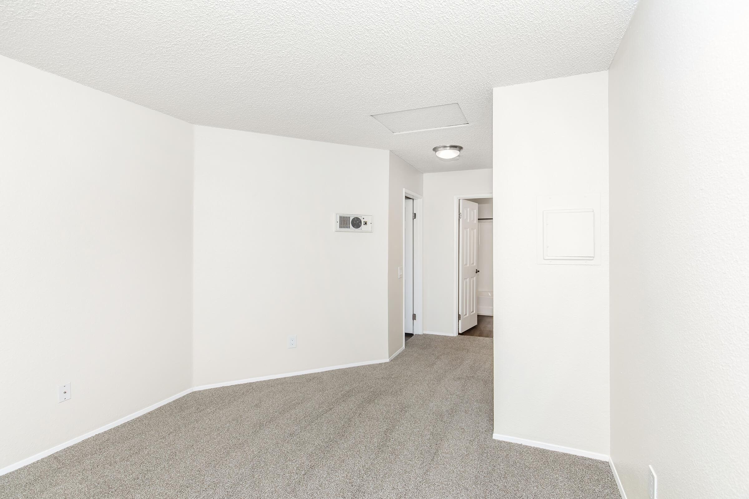 An empty room with light beige carpet and white textured walls. The corner shows a wall-mounted air conditioning unit. A doorway leads to a bathroom, visible in the background, while another door is partially open, suggesting additional space or storage. Natural light enhances the bright, clean atmosphere.