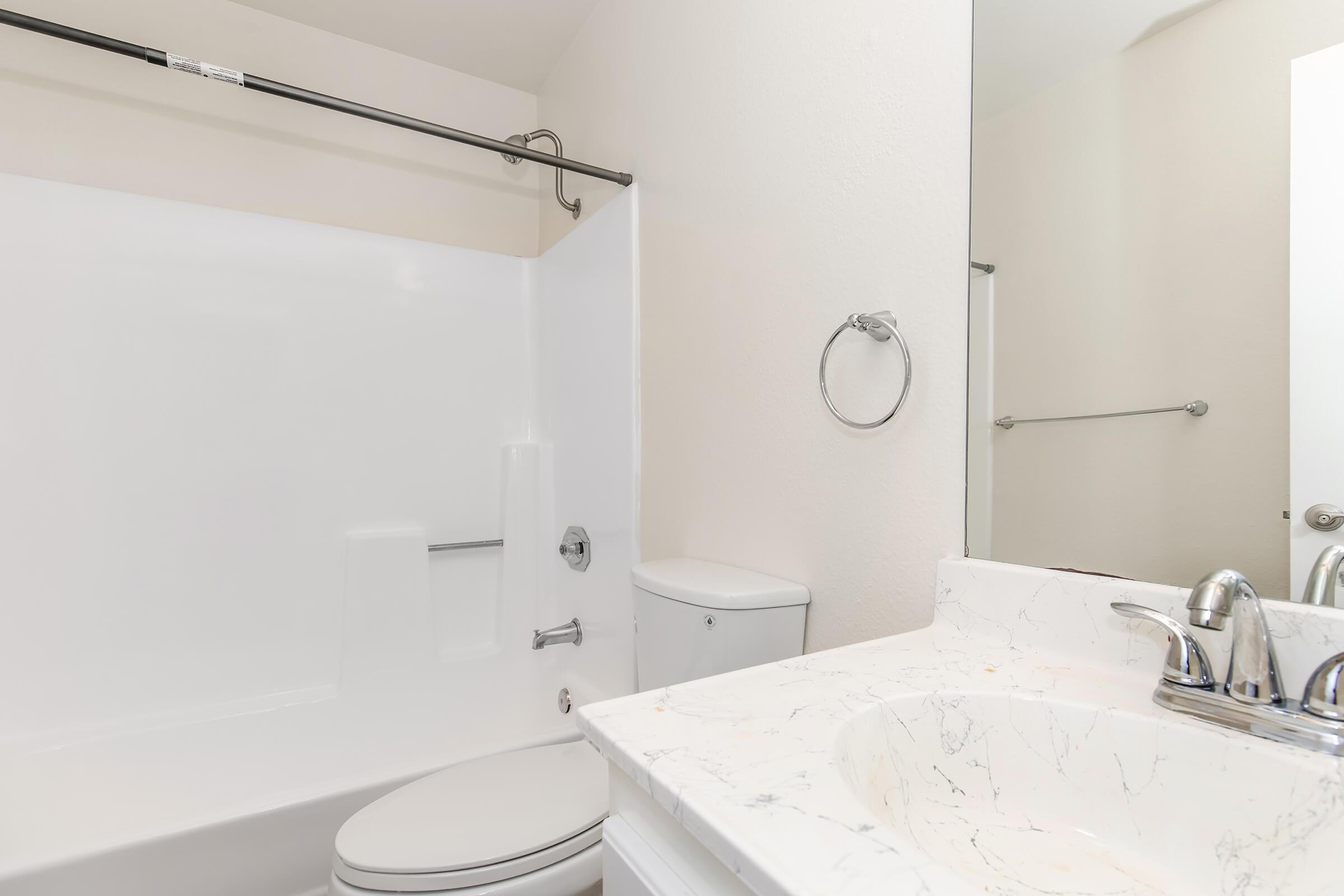 A clean and modern bathroom featuring a white bathtub with a shower, a toilet, and a marble countertop sink. Bright lighting reflects off the surfaces, creating a fresh and spacious appearance. Neutral-colored walls enhance the overall aesthetic of the space.