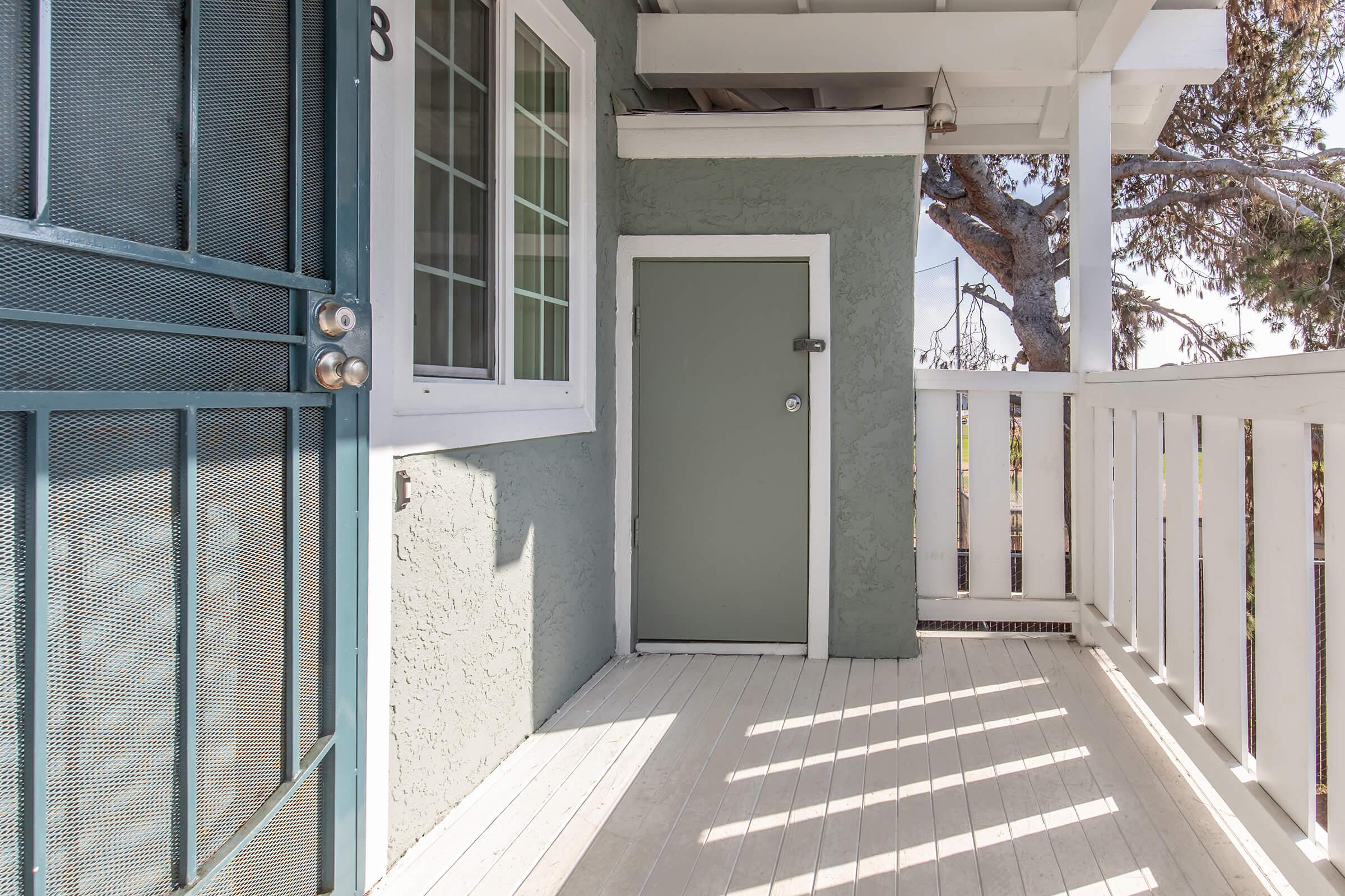 A small outdoor patio area with a light green door and a white railing. The floor is light wood, and there's a blue screen door visible. Sunlight is casting shadows on the floor, and a tree is partially visible in the background. The walls are painted a light gray color.