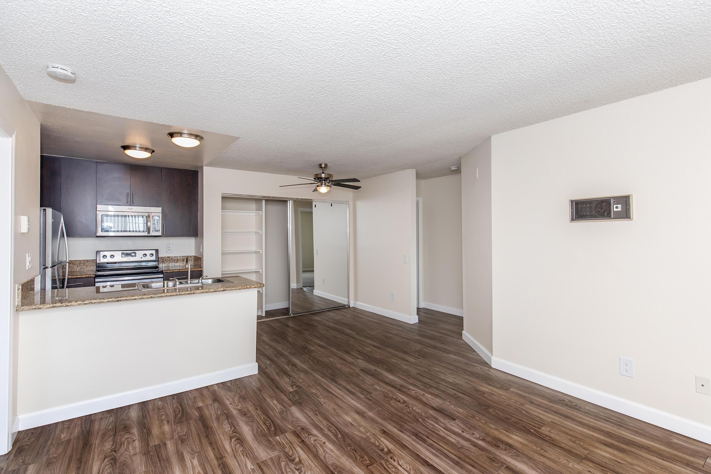 A well-lit, modern apartment interior featuring a kitchen with stainless steel appliances, a granite countertop, and an open layout leading to a living area with a ceiling fan. The space has neutral wall colors and a laminate wood floor, with a closet and doorway leading to another room.