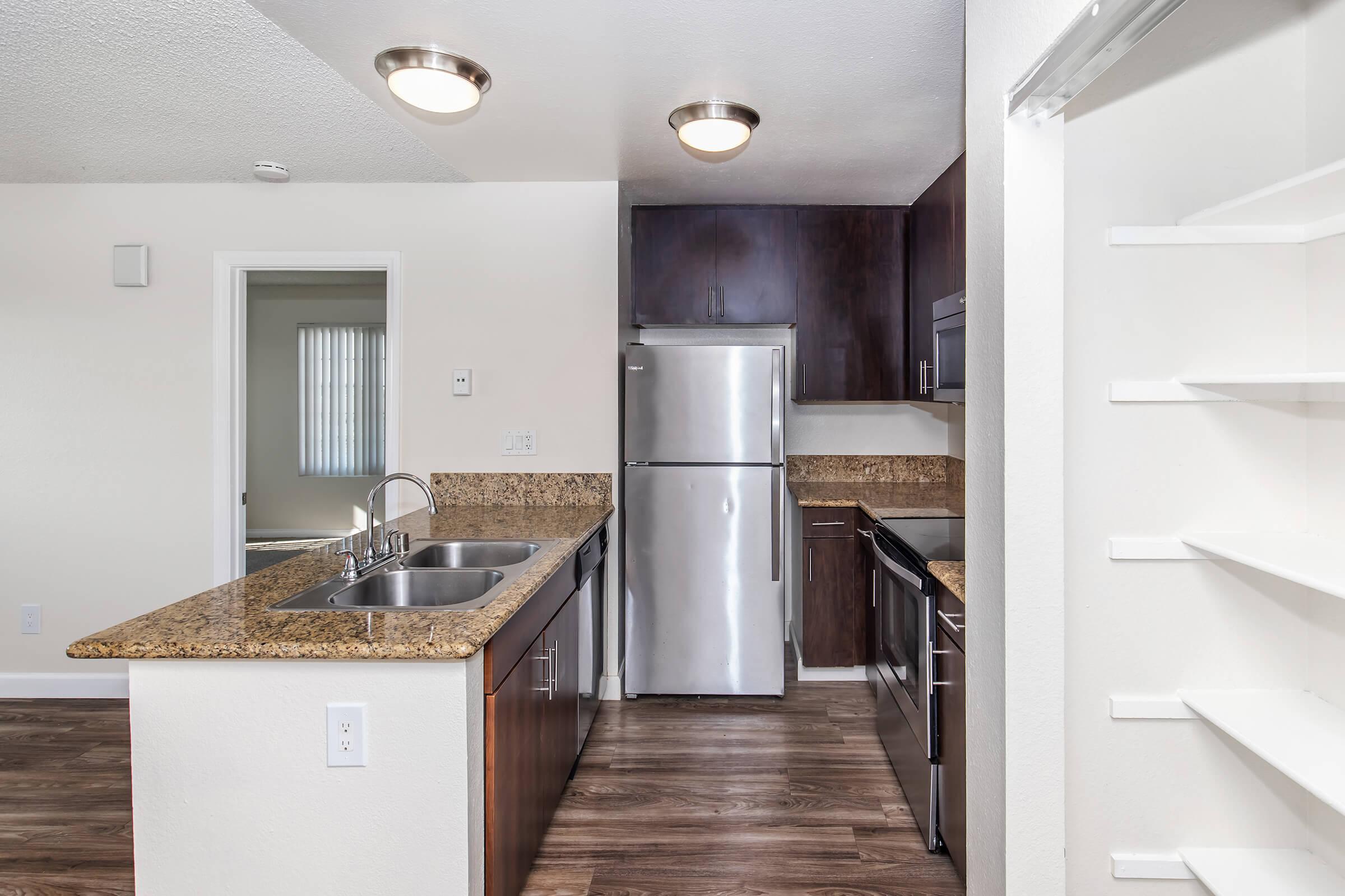 Modern kitchen featuring dark wooden cabinetry, stainless steel appliances including a refrigerator and dishwasher, and granite countertops. The space includes a large sink and an open layout with natural light coming from a nearby window.