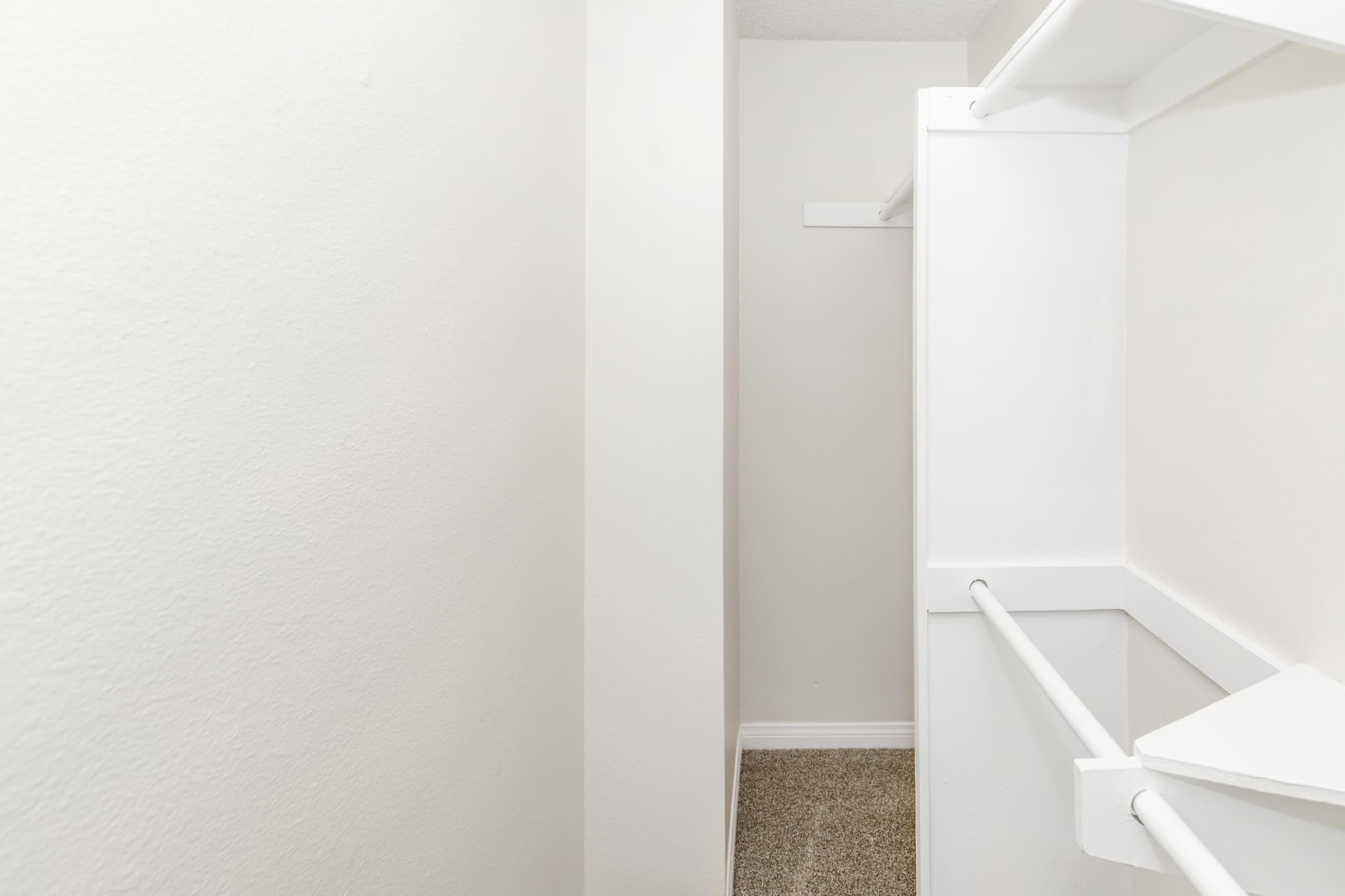 A clean, empty closet with light-colored walls and a carpeted floor. The closet features a white hanging rod and shelving. The space is minimalistic and well-lit, giving an organized appearance.